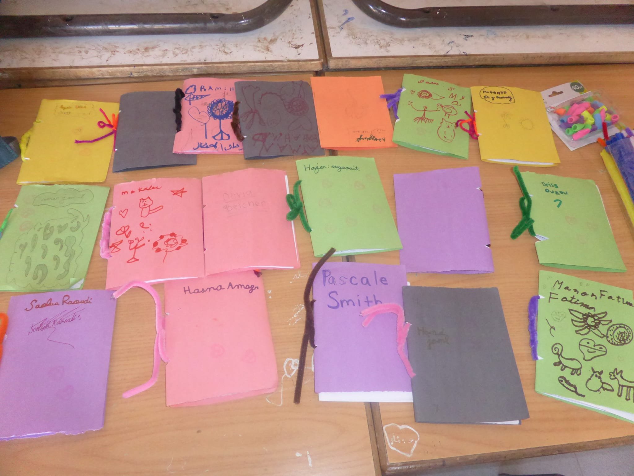 Colorful folded papers with handwritten names and drawings on a wooden surface, along with small bags of colored beads or clips.