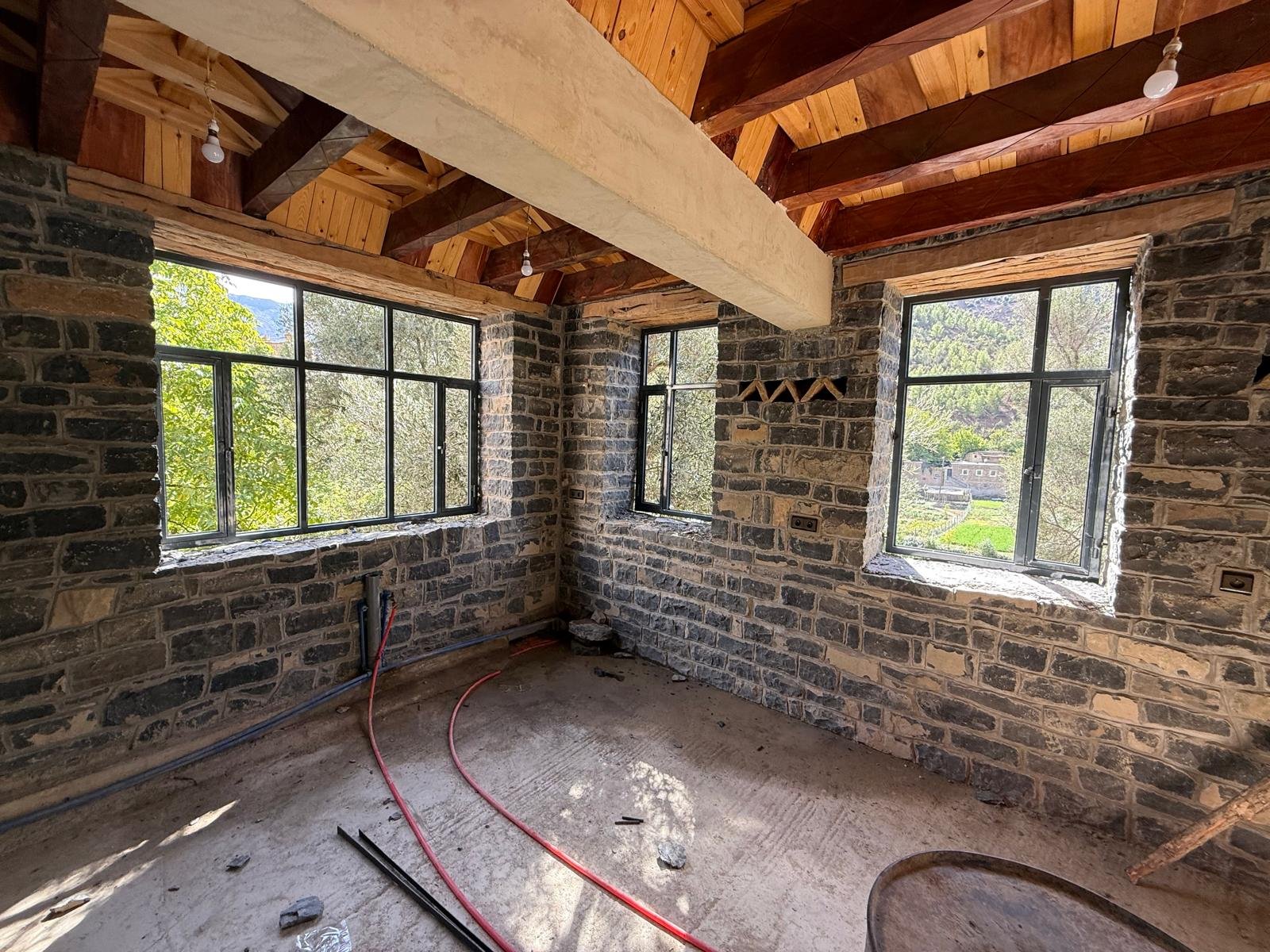 The interior of a room under construction with stone brick walls, large windows, and a wooden ceiling with exposed beams.