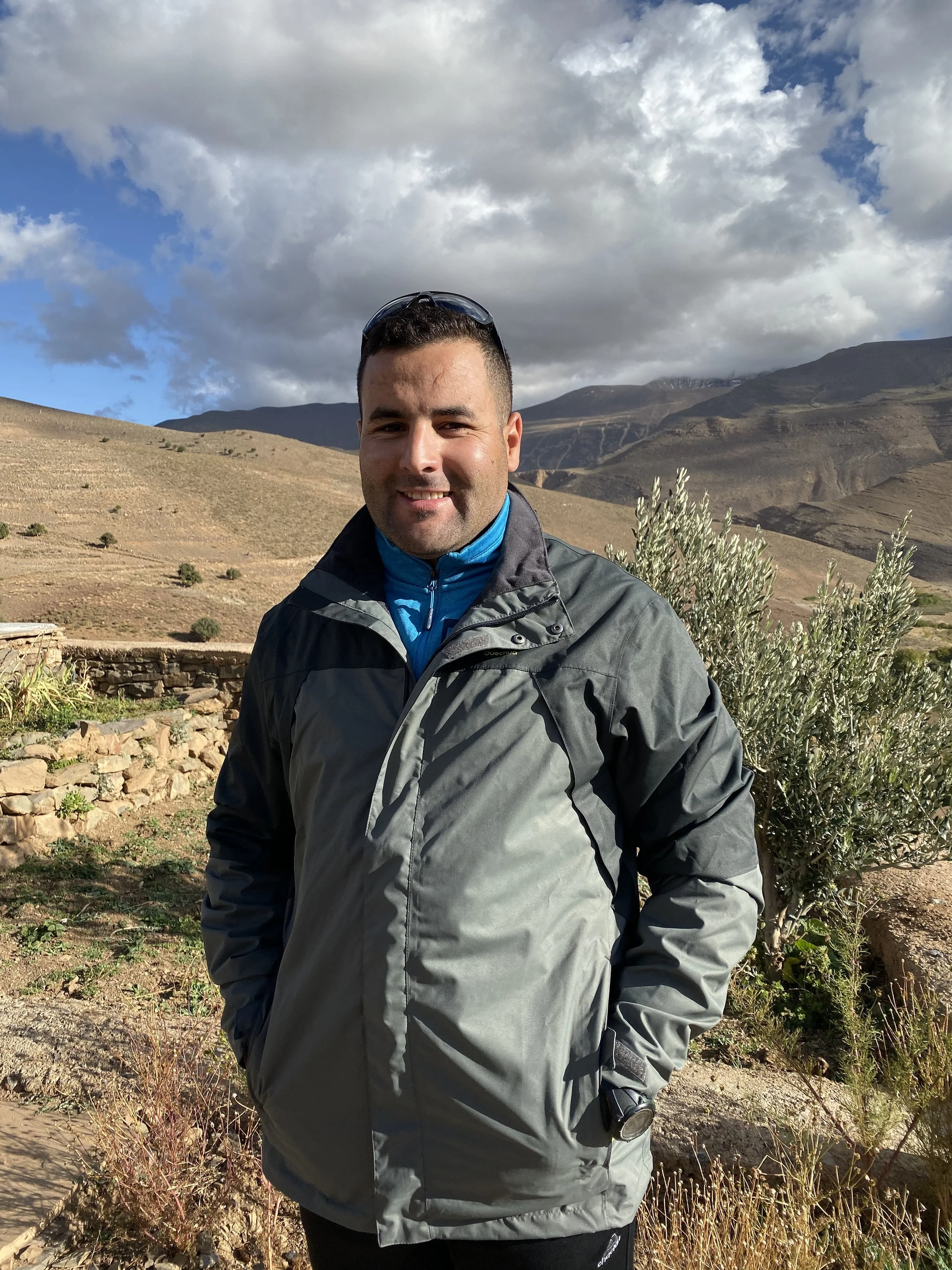 A man in outdoor gear smiling in a mountainous landscape with partly cloudy sky, dry grass, and shrubs.