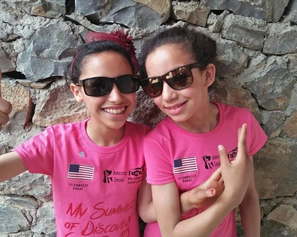 Two young girls wearing pink shirts and sunglasses posing in front of a stone wall, smiling and making playful hand gestures.