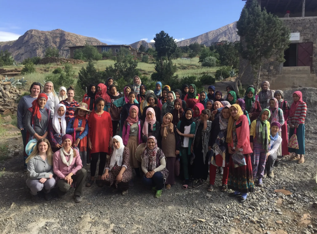 Group of women and girls posing outdoors in a mountainous area with greenery and buildings in the background.