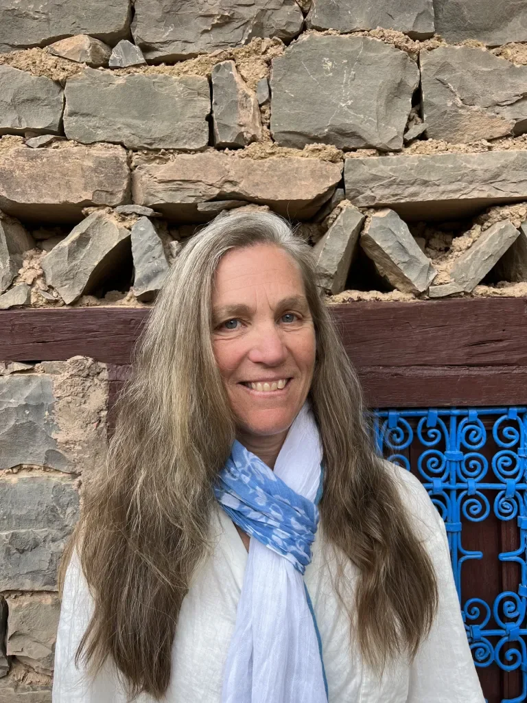 A woman with long, wavy, gray hair smiling in front of a stone wall with a decorative blue wrought iron gate.