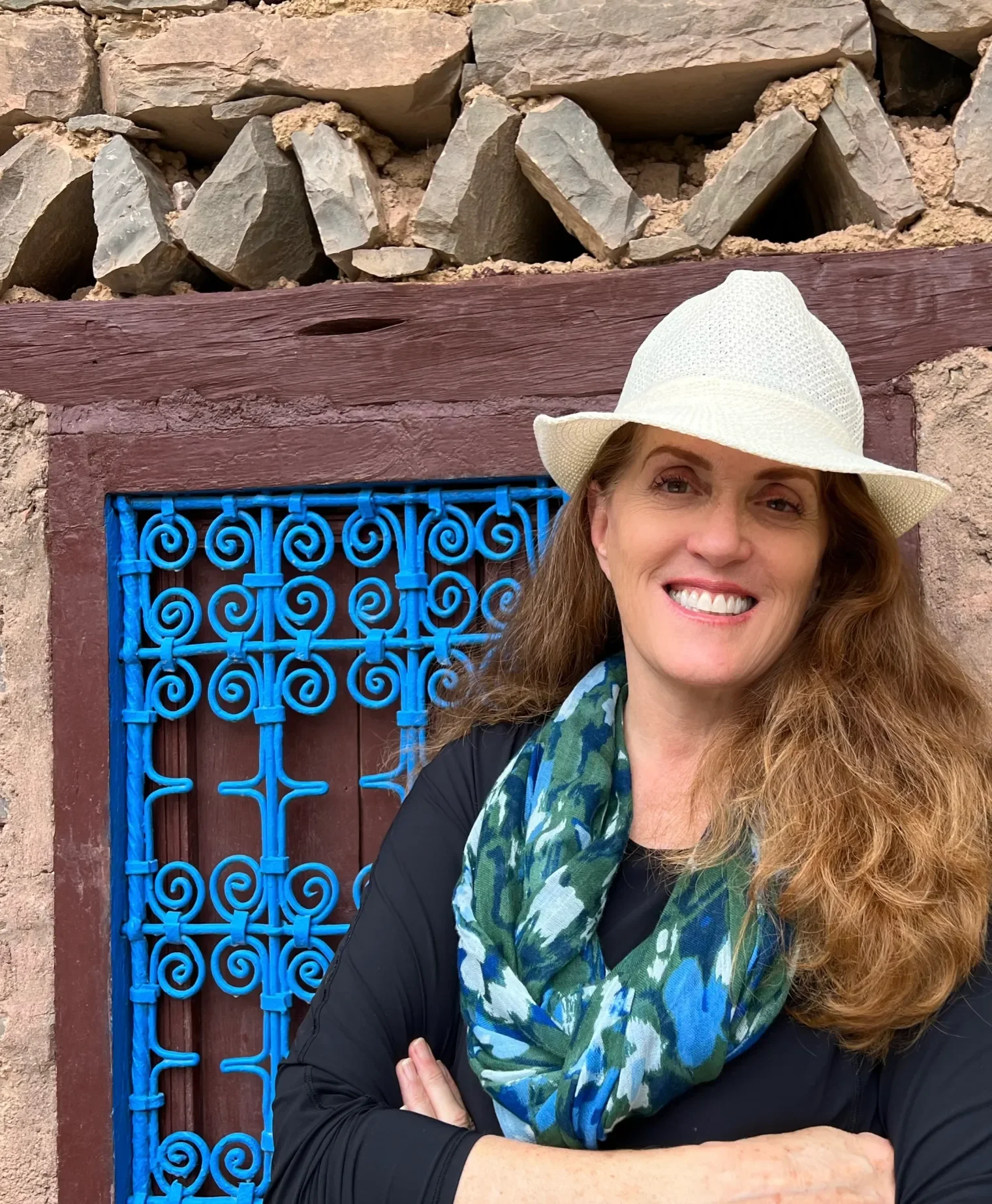 Woman with long curly hair wearing a white hat and a green and blue patterned scarf, smiling and crossing her arms, standing in front of a stone wall with a wooden structural element and a blue decorative iron grille.
