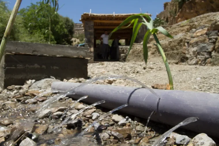 A large gray pipeline releasing water onto a rocky ground with a small plant nearby. In the background, a wooden shelter with two people inside, set in a rural area with a stone hill and trees.