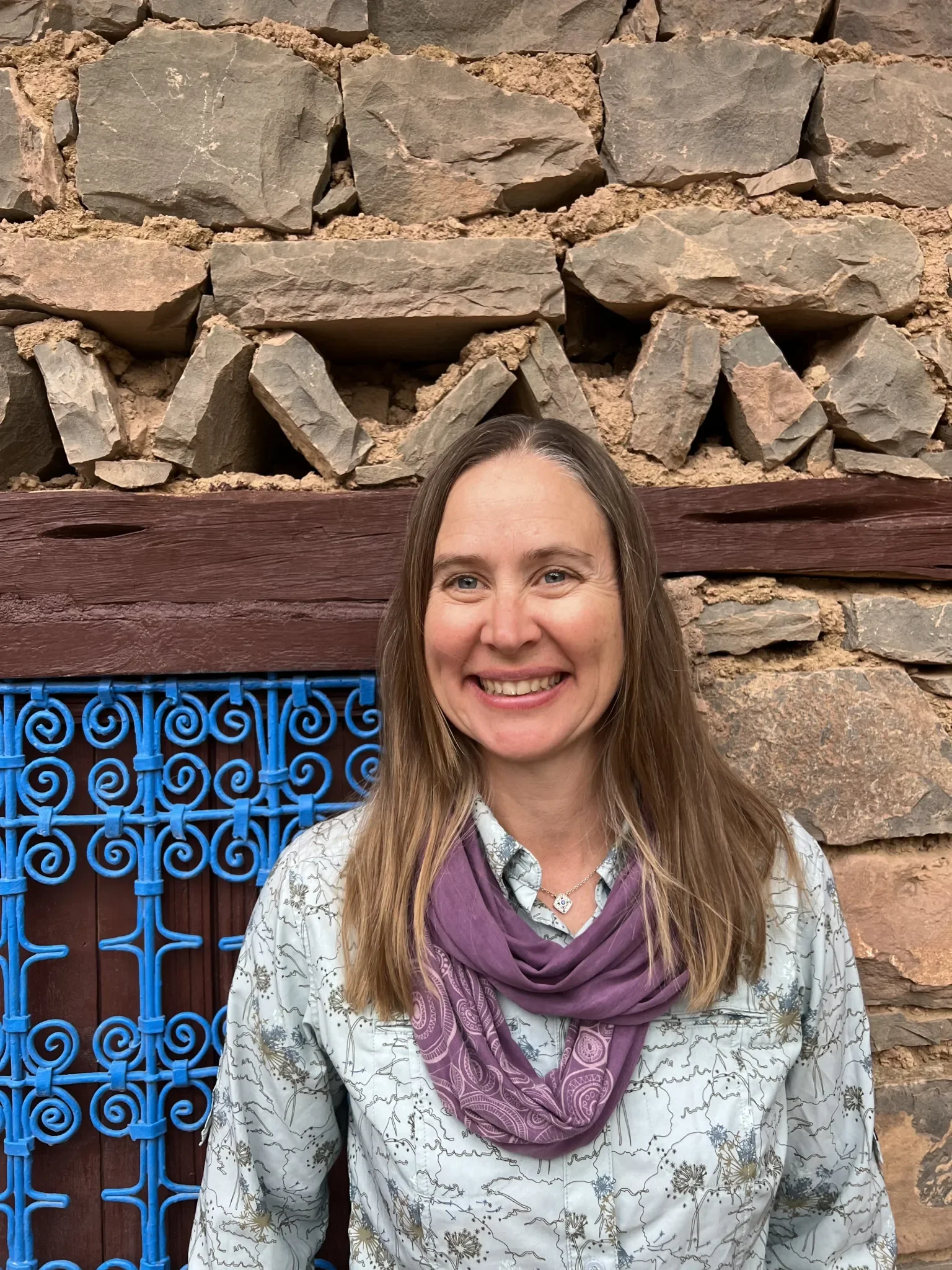 A smiling woman with long brown hair standing in front of a rock wall and a decorative blue metal fence.