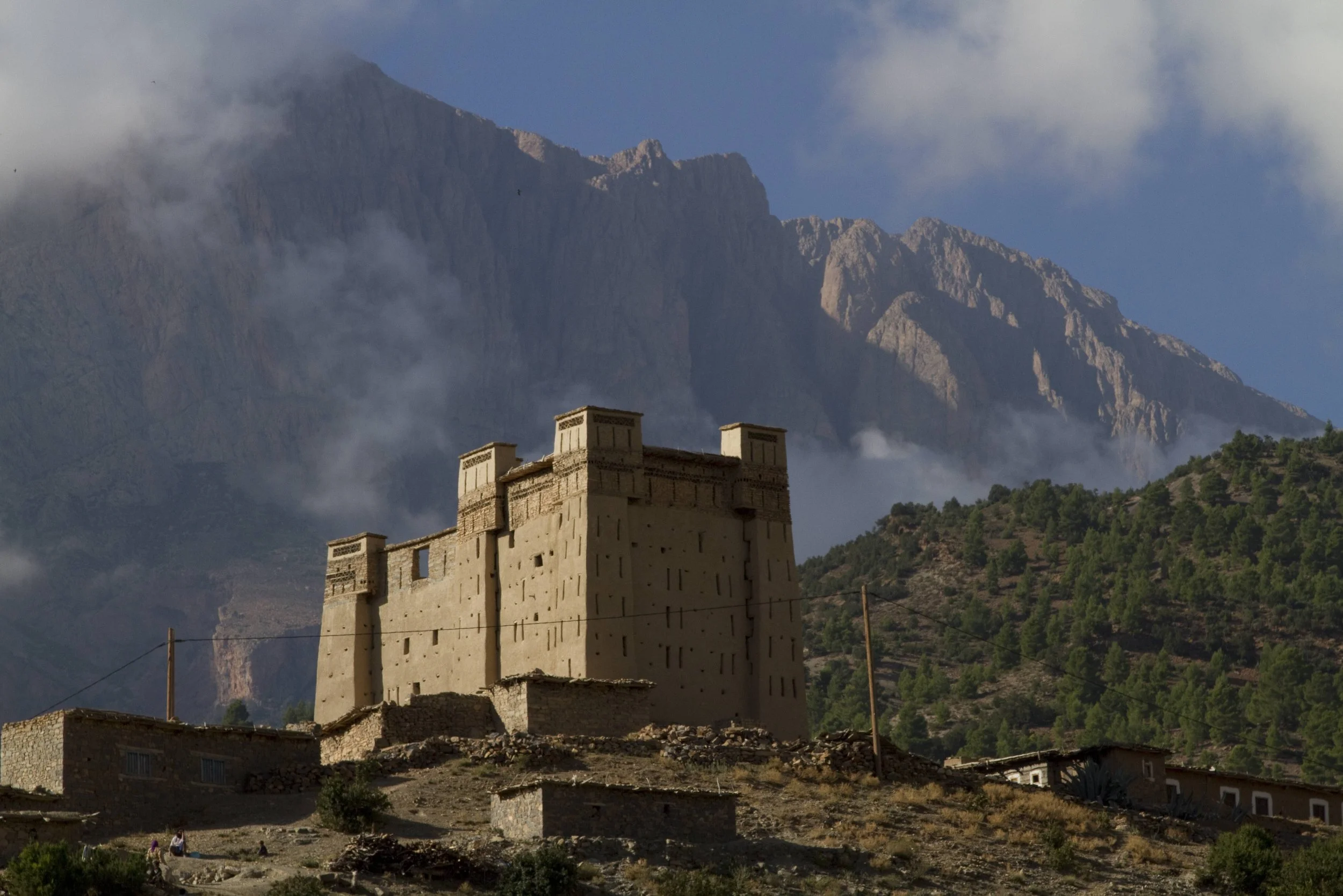A mountain landscape with a historic building in the foreground, surrounded by trees and smaller houses, with tall mountains and clouds in the background.