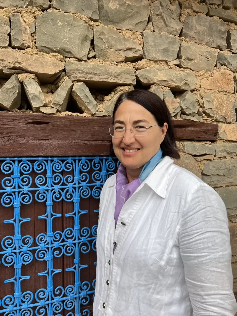 A woman with glasses and dark hair, smiling, standing next to a blue wrought-iron gate in front of a stone wall.