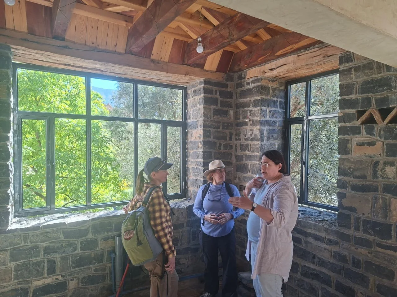 Three women having a conversation inside a room with stone walls and large windows showing green trees outside. One woman is gesturing while holding a water bottle.