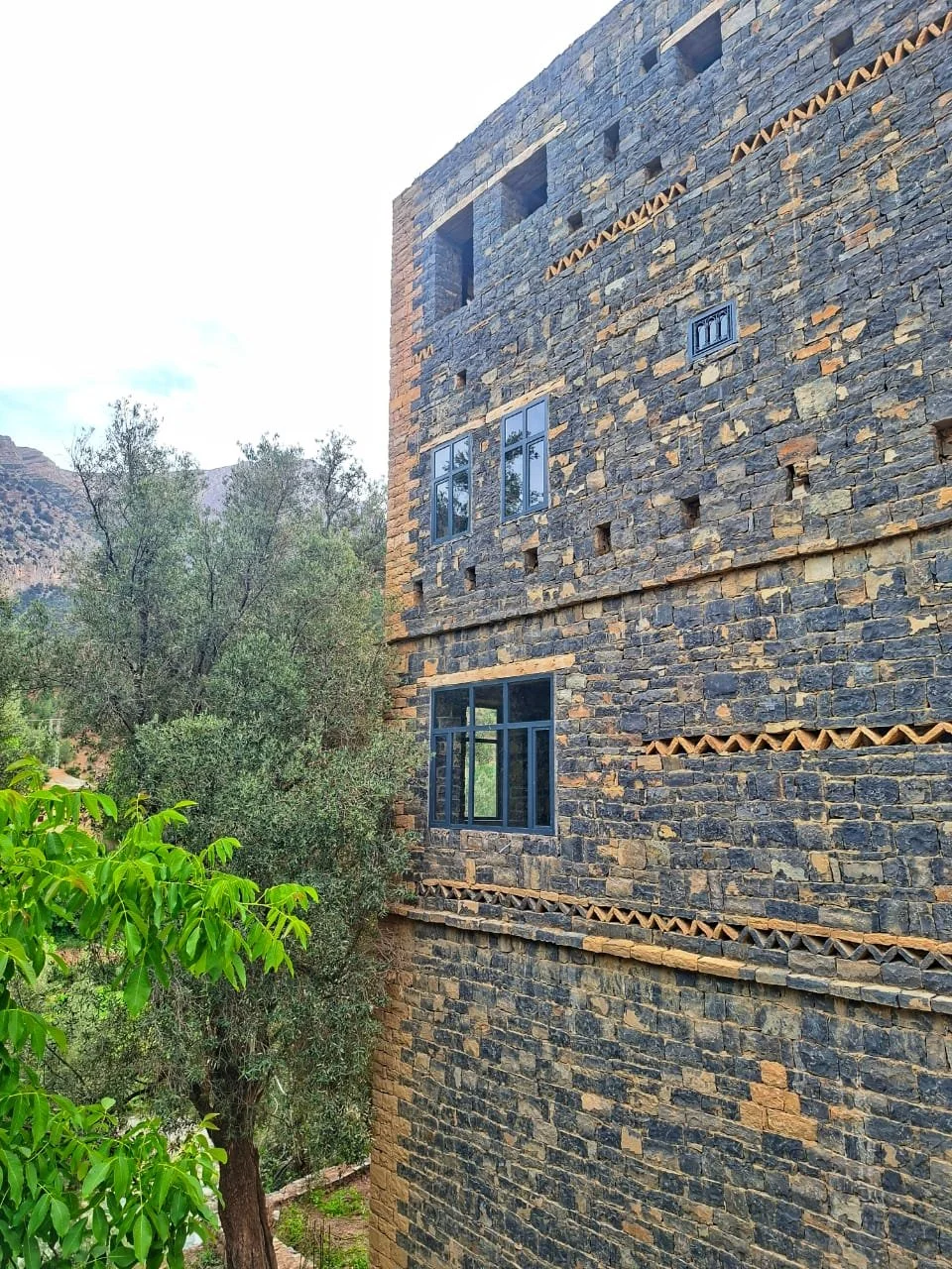 A multi-story building with a stone facade surrounded by trees and mountains in the background.