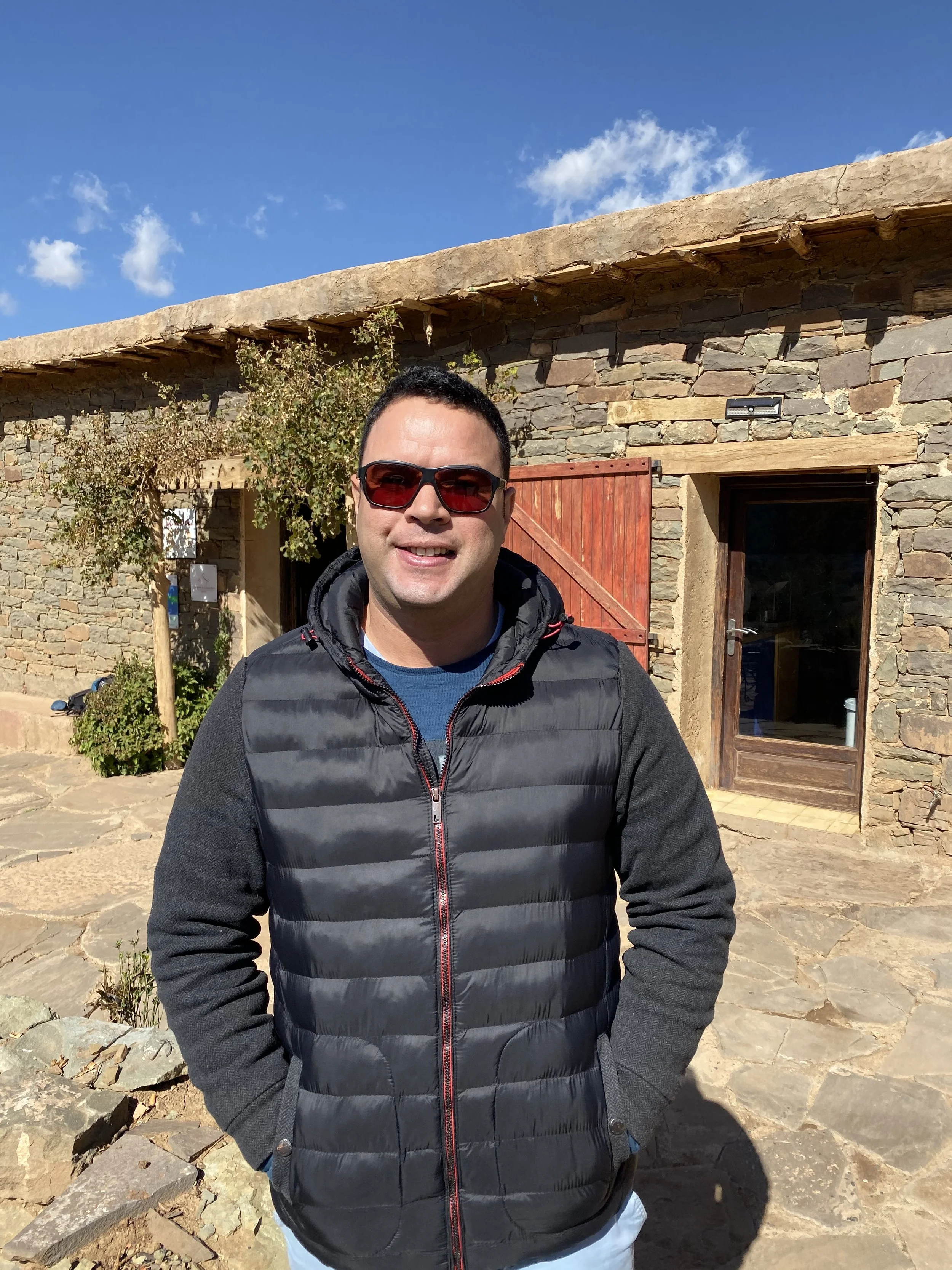 A man wearing sunglasses and a black puffer jacket smiling outdoors in front of a stone building with a red door and a wooden beam over the doorway, with a blue sky and some scattered clouds above.