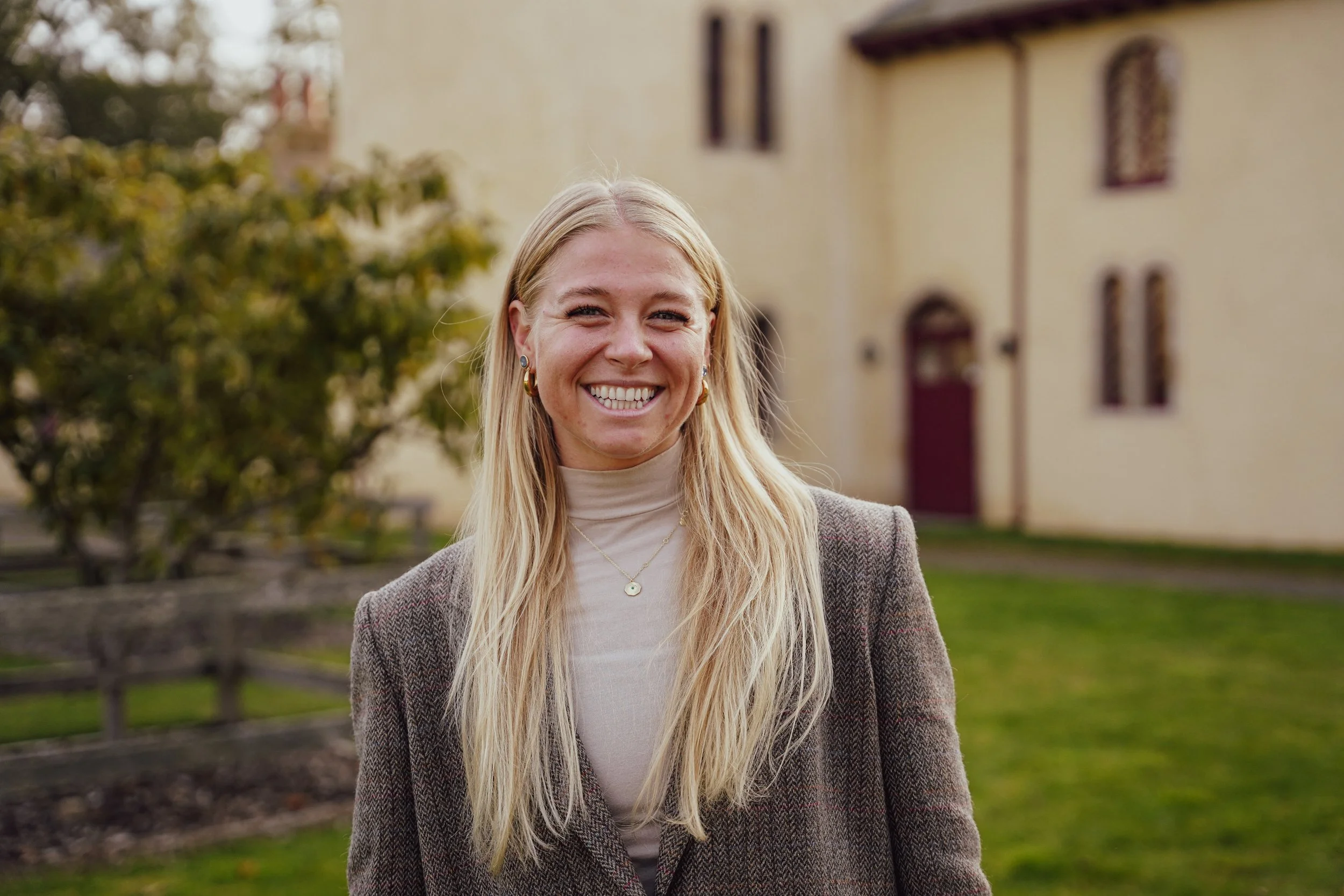 Young woman with long blonde hair smiling outdoors, wearing earrings, a beige turtleneck, and a plaid blazer, in front of a building and greenery in Moray, Scotland.