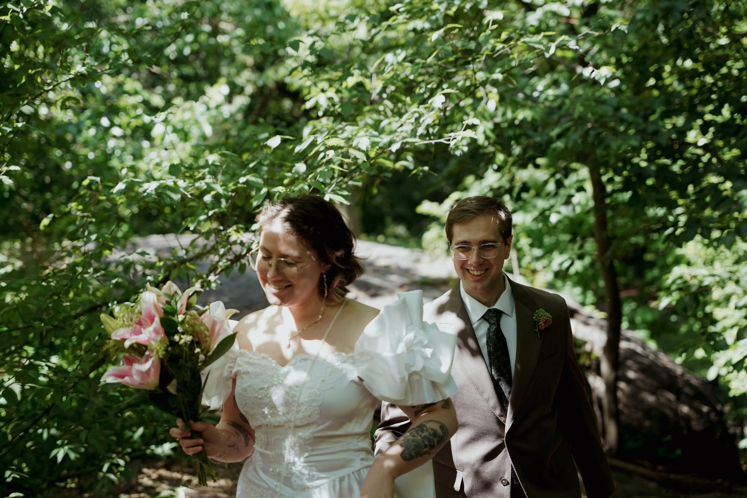 A bride and groom smiling and walking outdoors under green trees, with the bride holding a bouquet of pink lilies.