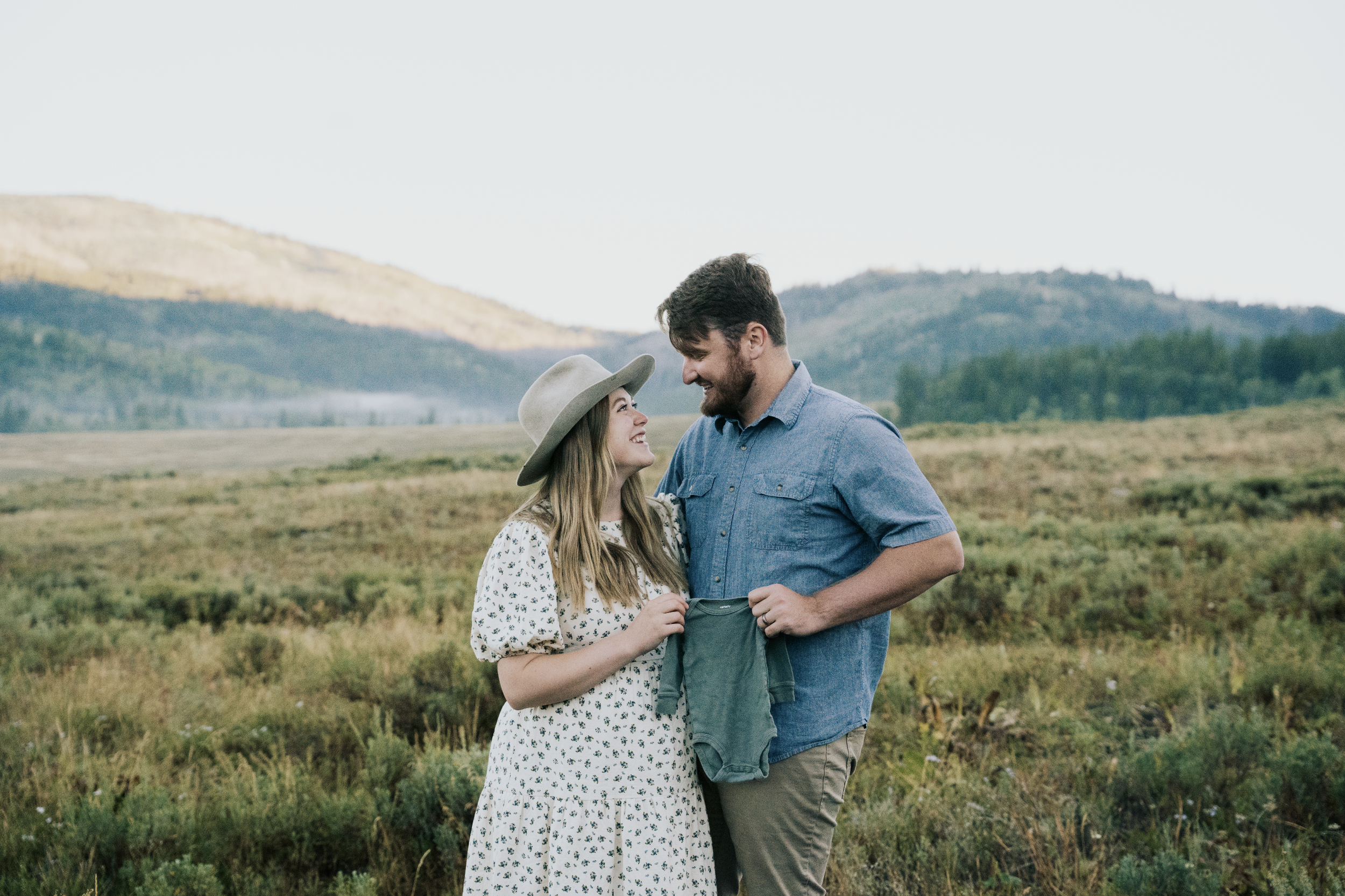 A young couple smiling and looking at each other, standing in a field with green grass and hills in the background.