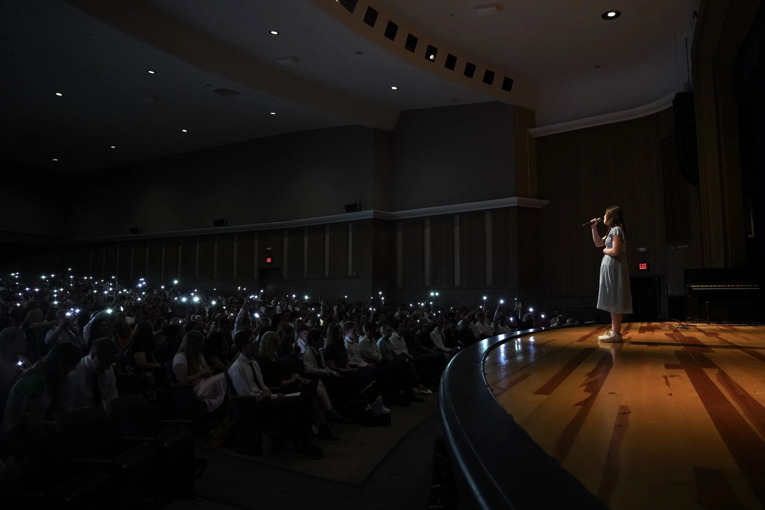 A girl standing on stage holding a microphone, facing an audience with numerous cell phone lights turned on, in an auditorium with a high ceiling and wooden wall panels.