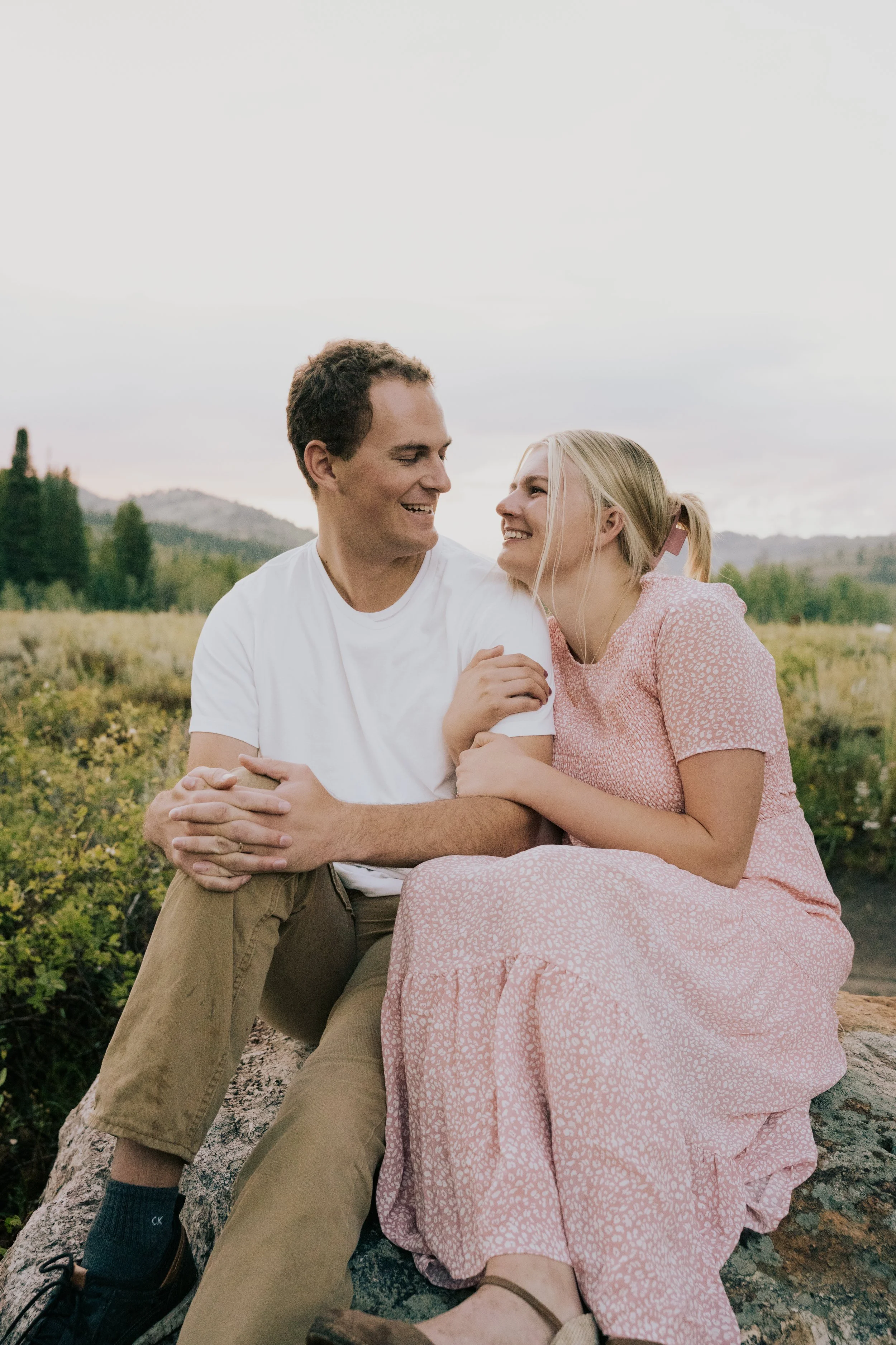 A happy couple sitting outdoors on a log in a field, smiling and looking at each other, with mountains and trees in the background during daytime.