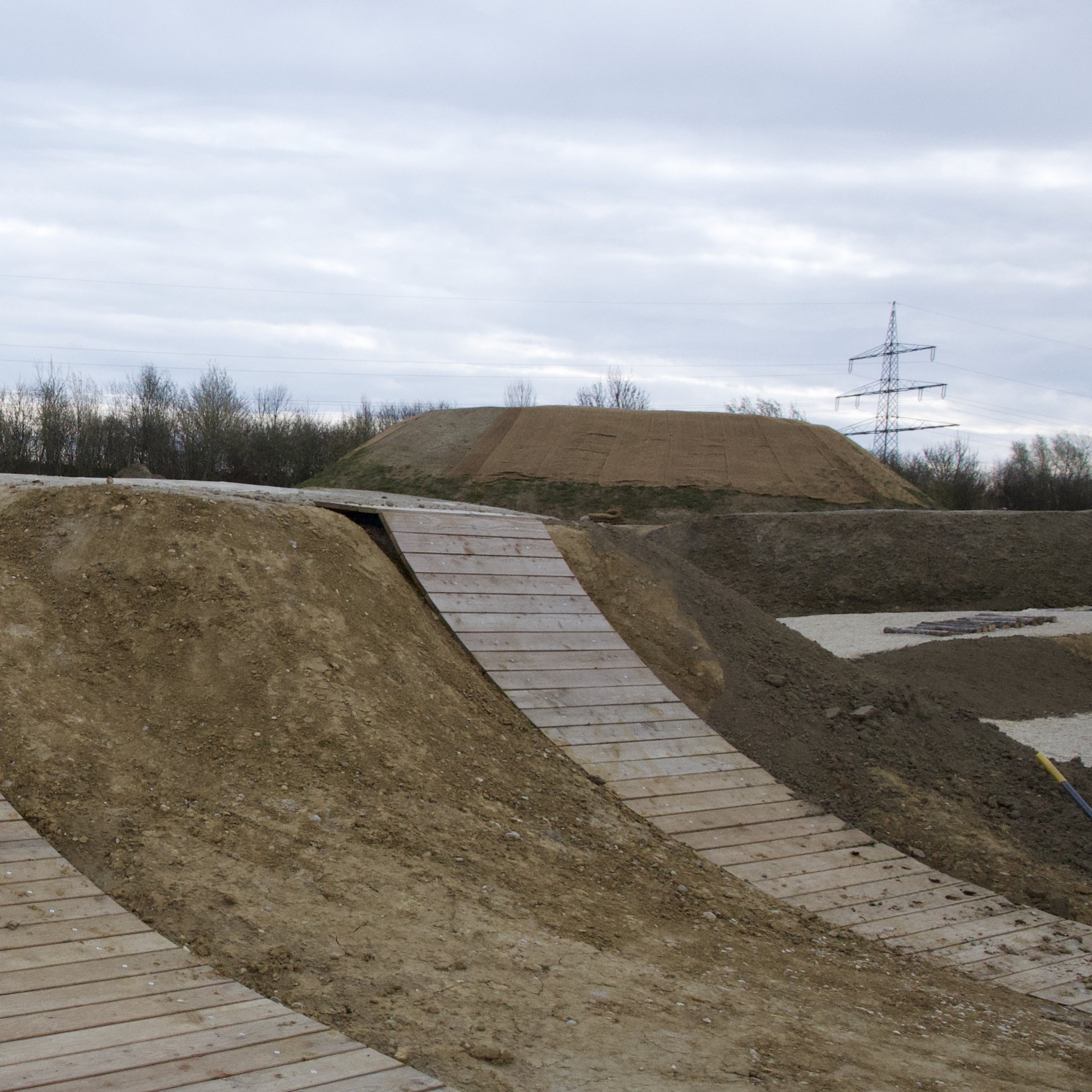 Der Bau einer BMX-Sprungstrecke mit Erdaufschüttungen und Holzrampe unter einem bewölkten Himmel, im Hintergrund sind Bäume und ein Strommast sichtbar.