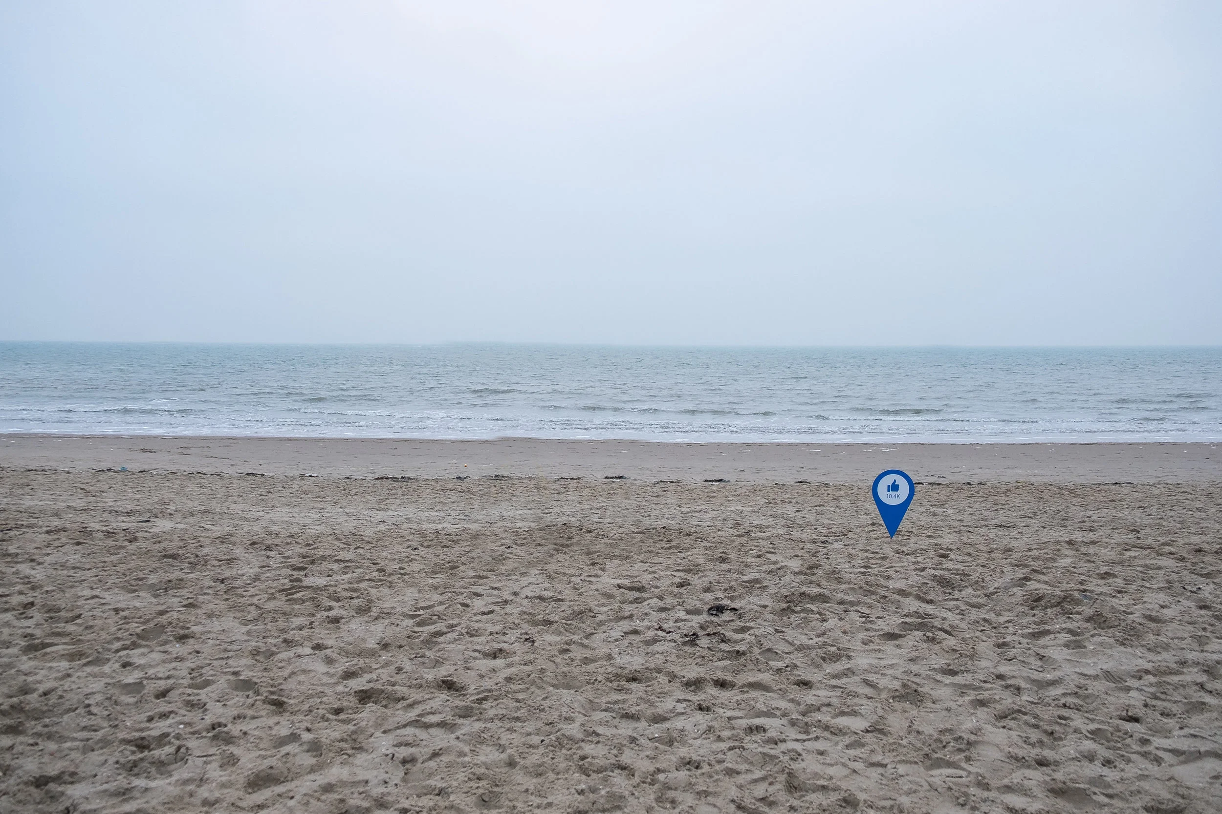 Empty beach with sand, ocean, and cloudy sky; a blue location marker with a thumbs-up icon and '10.4K' is placed on the sand.