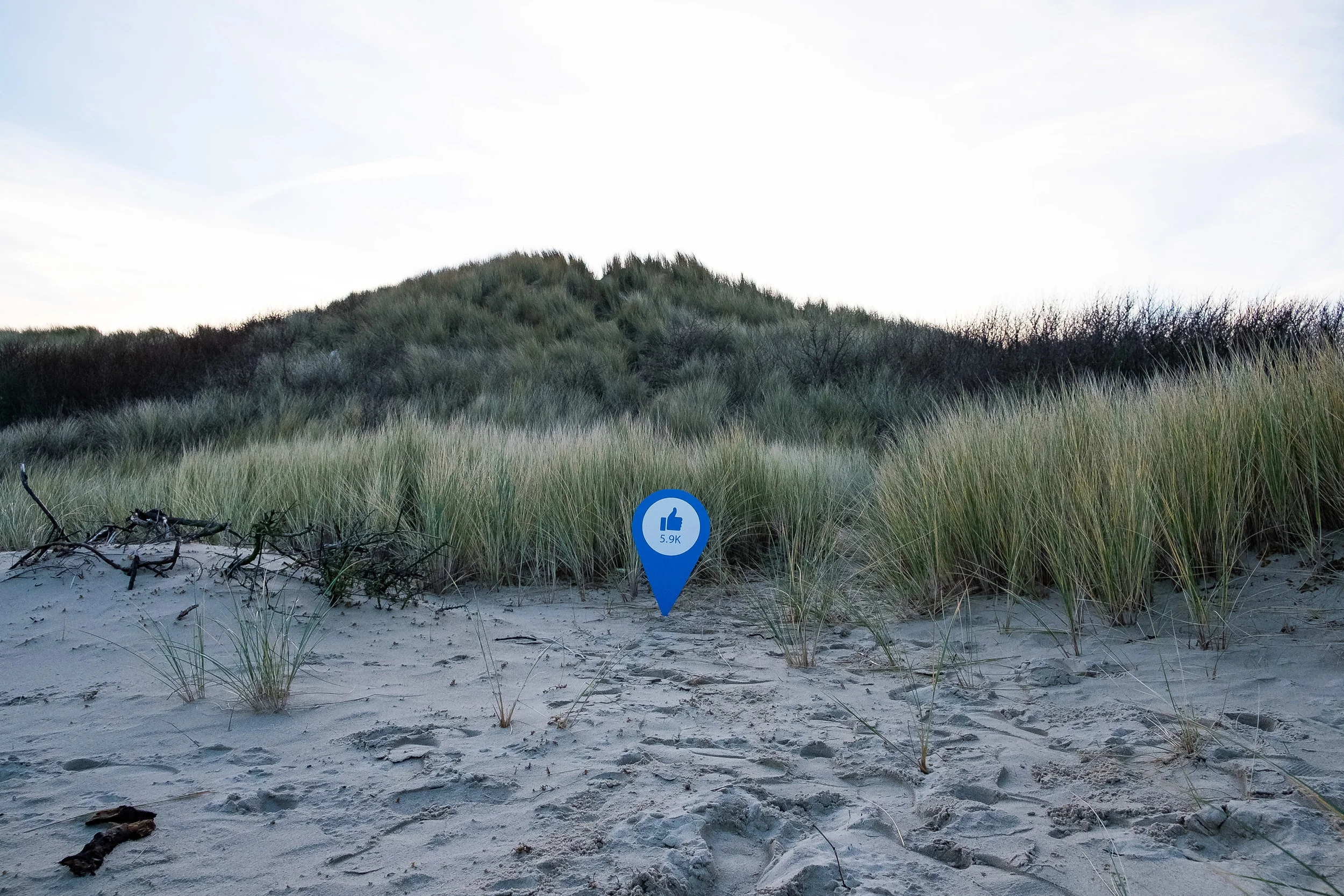 A sandy beach with tall grass and shrubs, with a hill in the background under a partly cloudy sky. A blue social media-like marker with a thumbs-up icon and '5.9K' likes stands in the foreground.