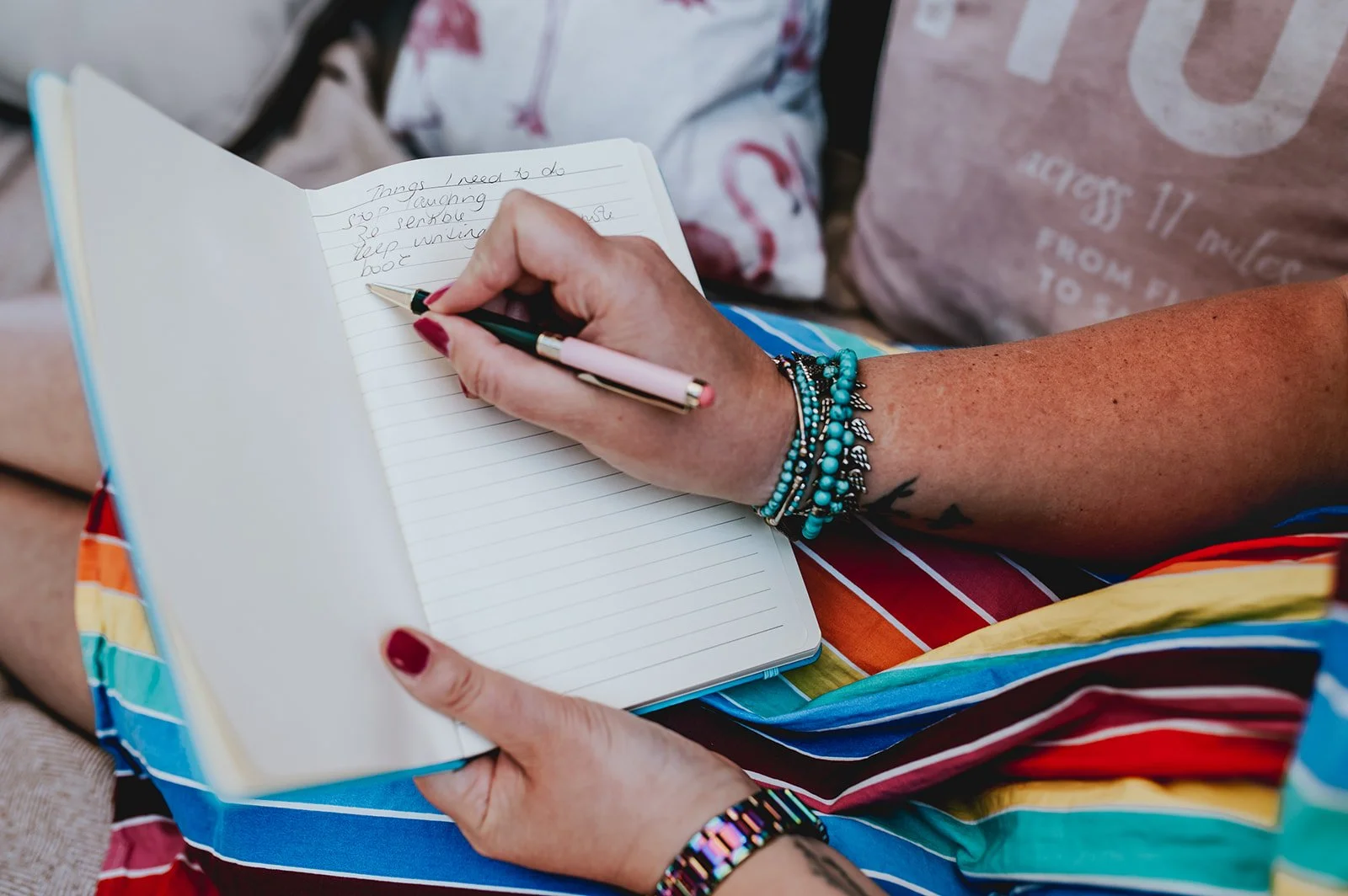 A woman is sitting outdoors, writing in a notebook with a black and pink pen. She has multiple turquoise and silver bracelets on her wrist and is wearing a colorful, striped skirt with rainbow colors. The background shows people wearing casual clothing.