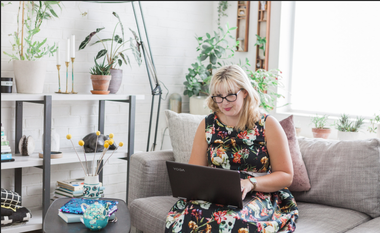 Beckie wearing glasses sitting on a couch with a laptop in a bright, plant-filled living room.