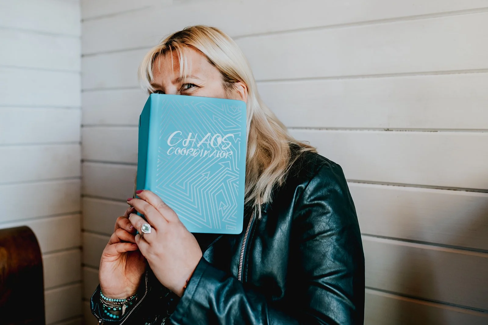 Beckie with blonde hair, wearing a black leather jacket, holds a blue book titled 'Chaos Coordinator' in front of her face, partially covering it, with a white paneled wall background.