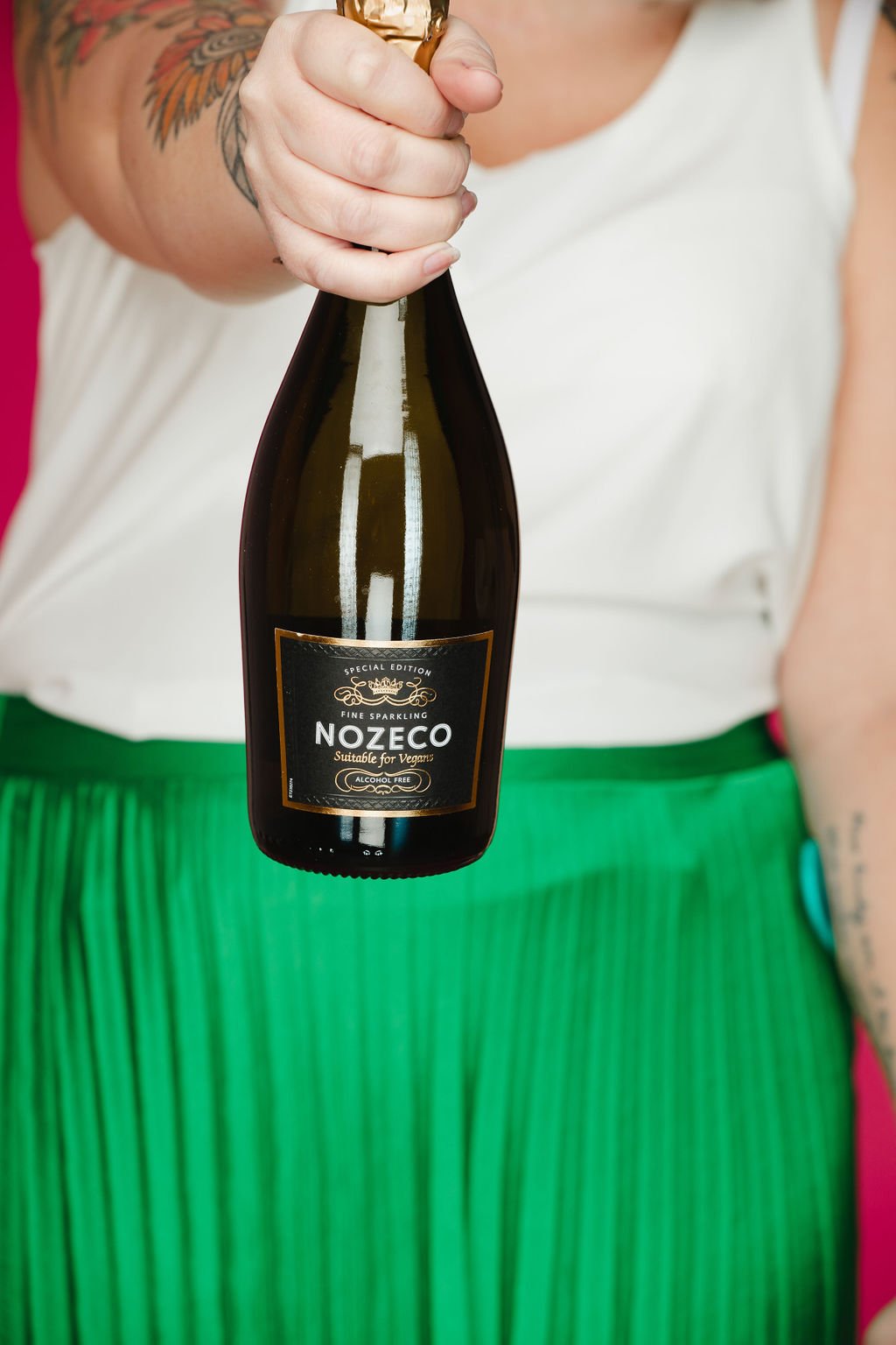 Beckie holding a bottle of NozeCo sparkling beverage with a black label, white text, and gold accents, wearing a white top and green pleated skirt, standing against a pink background.