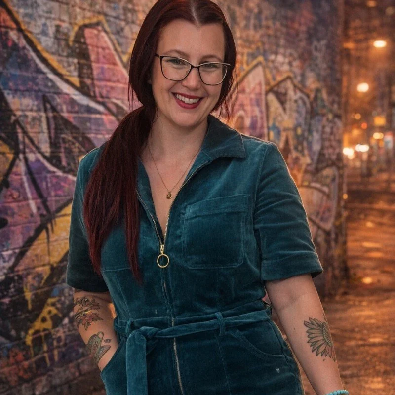 Beckie with long red hair and glasses, smiling, wearing a teal velvety jumpsuit with a zipper, standing in front of a graffiti-covered brick wall at night.
