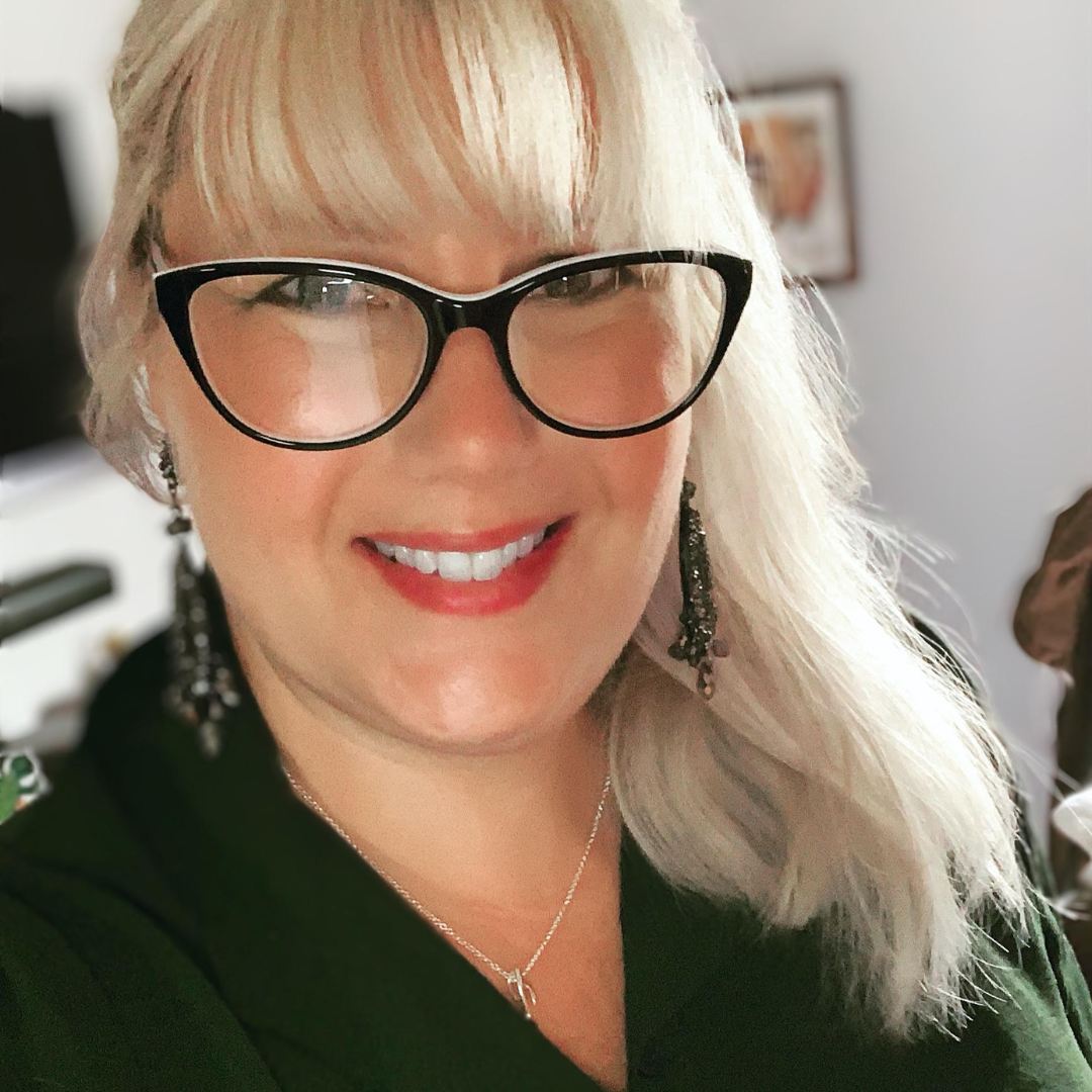 Beckie has blonde hair, wearing black glasses, earrings, a necklace, and a black outfit, smiling.