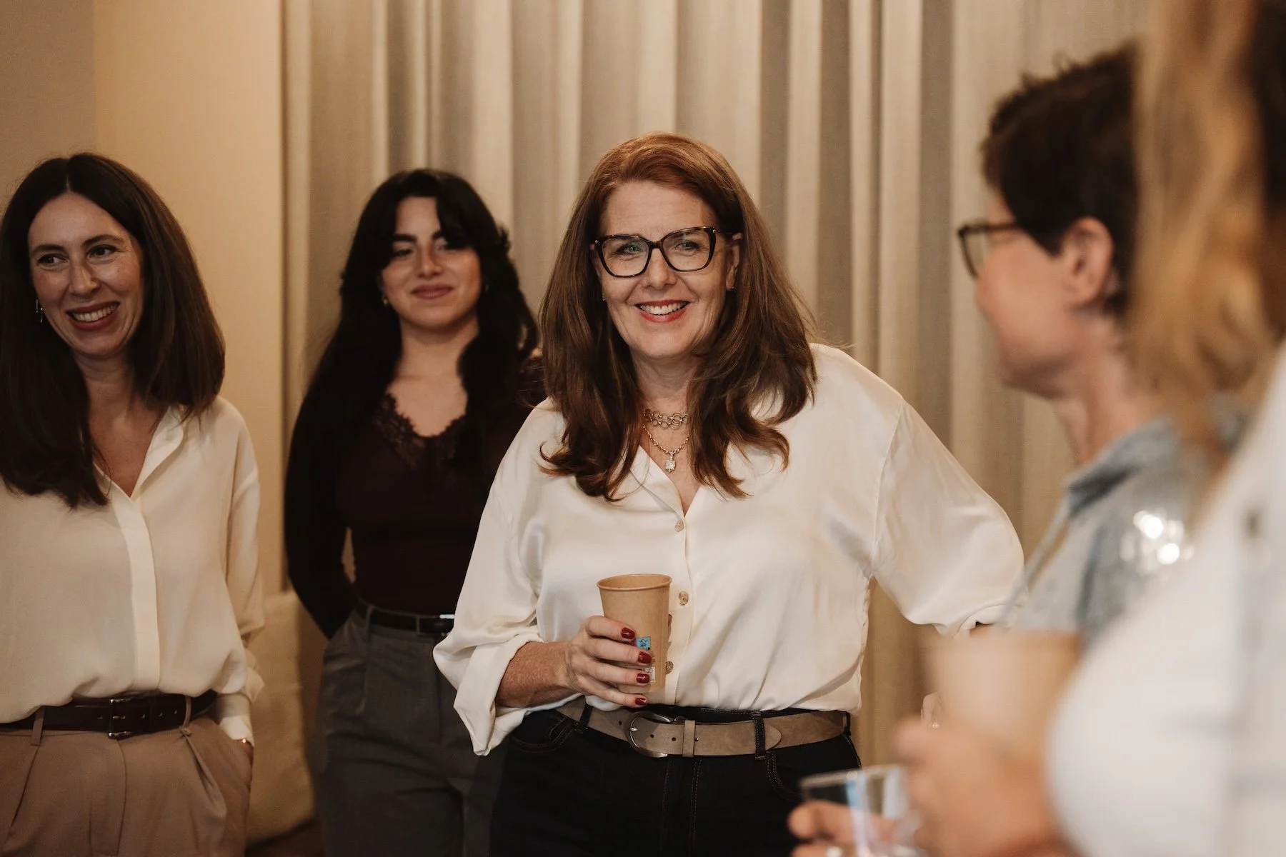 Eine Gruppe von Frauen im Gespräch bei einer gesellschaftlichen Veranstaltung, eine Frau trägt eine weiße Bluse, hält einen Pappbecher und lächelt.
