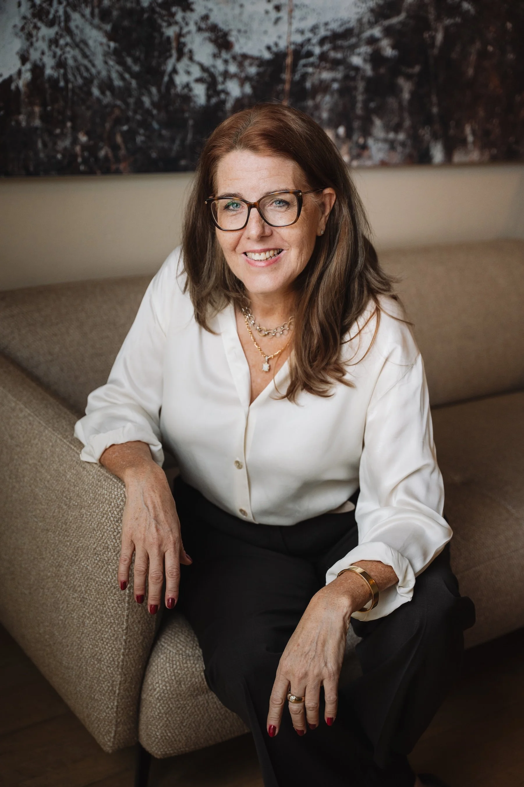 Eine lächelnde Frau mit braunen Haaren, Brille, weißen Bluse und dunklen Hosen sitzt entspannt auf einem Sofa in einem modernen Raum.