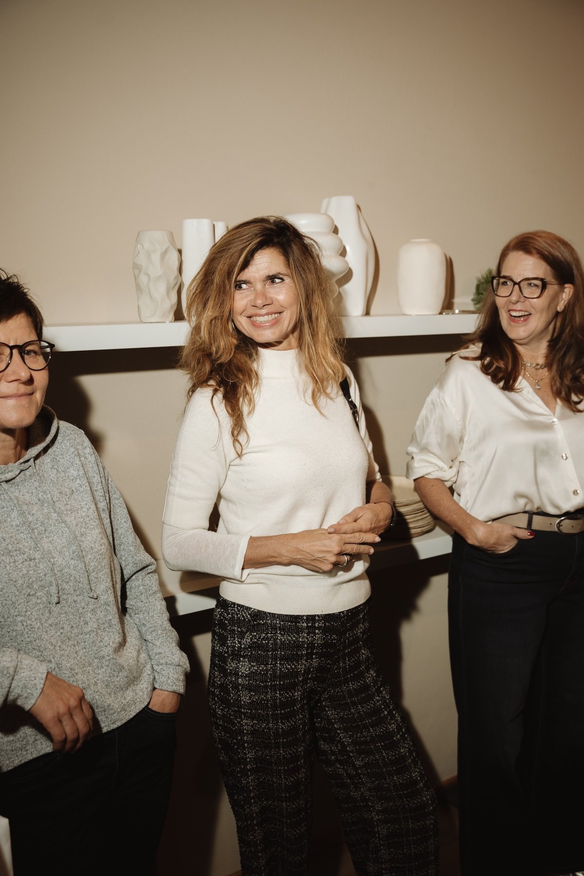 Drei Frauen, die in einem Raum stehen, lächeln und sich unterhalten, mit einer weißen Wand und Vasen im Hintergrund.