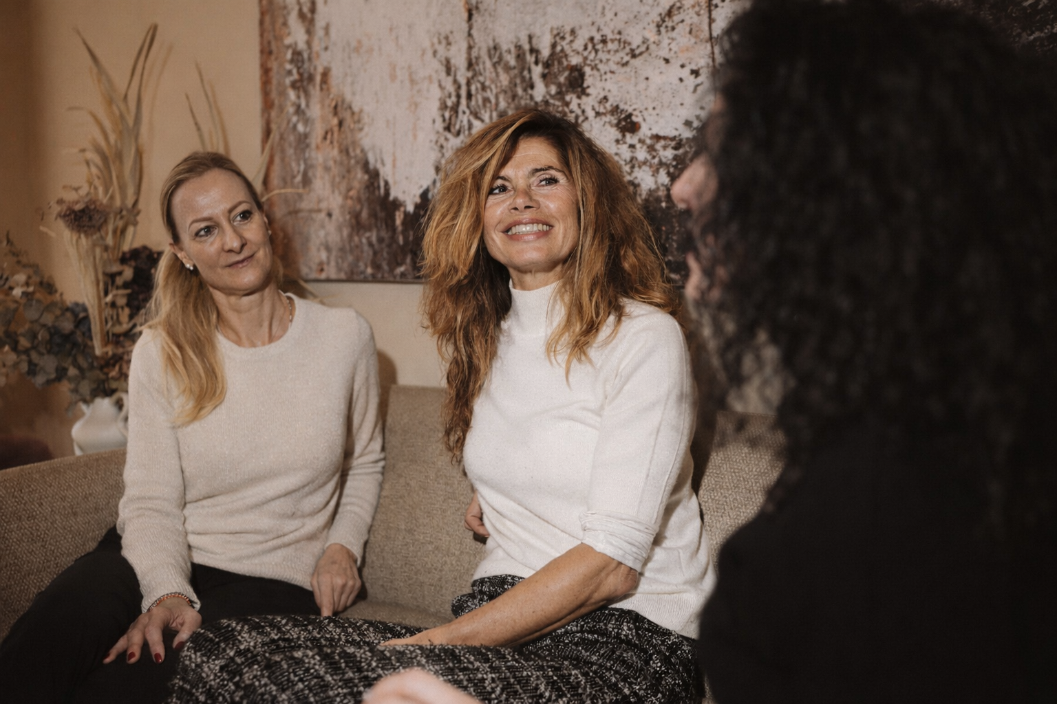 Drei Frauen sitzen in einem Wohnzimmer, eine lächelt und scheint im Gespräch zu sein, während die anderen beiden zuhören.