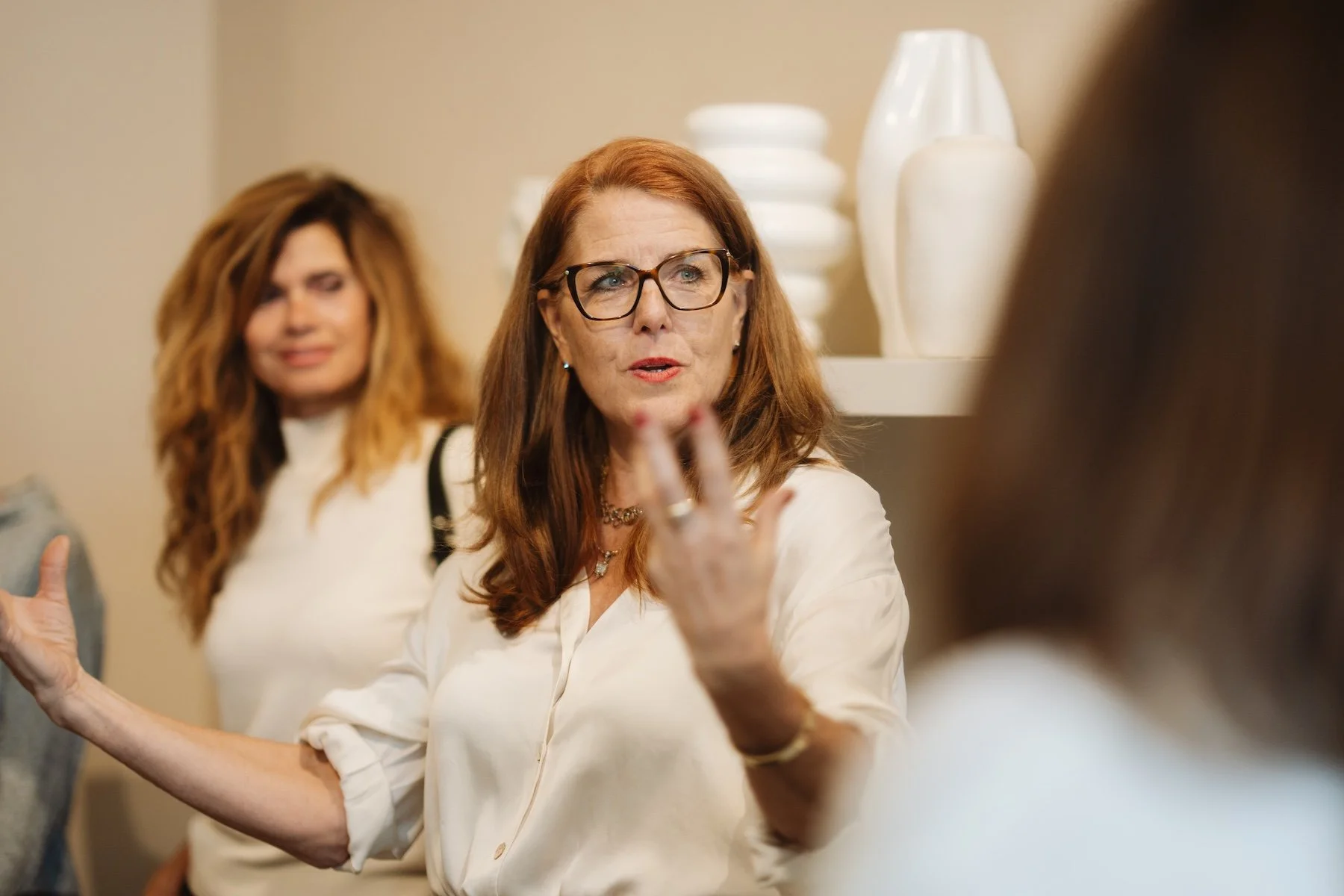 Drei Frauen im Gespräch, die zentrale Frau trägt Brille und eine weiße Bluse, die andere Frau im Hintergrund hat lange, lockige Haare und trägt ebenfalls eine weiße Kleidung.