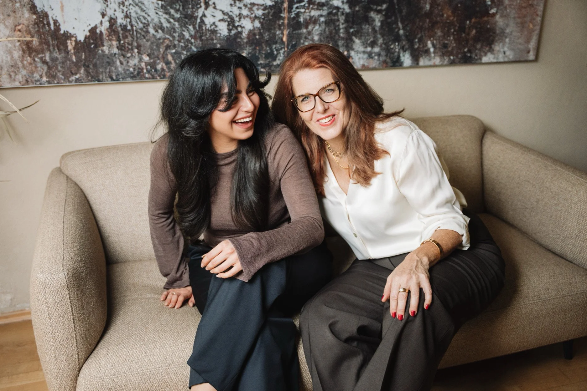 Zwei lachende Frauen sitzen gemeinsam auf einem Sofa, eine mit schwarzen Haaren und die andere mit roten Haaren und Brille, während sie bei einem gemütlichen Treffen lachen.