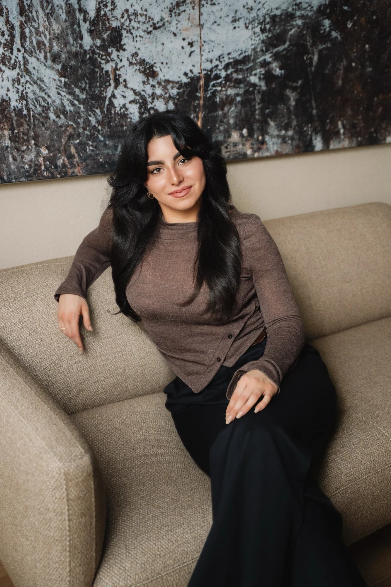 Eine junge Frau mit longem schwarzen Haar sitzt entspannt auf einem beige Couch, trägt ein braunes Oberteil und schwarze Hosen, im Hintergrund ein abstraktes Kunstwerk.