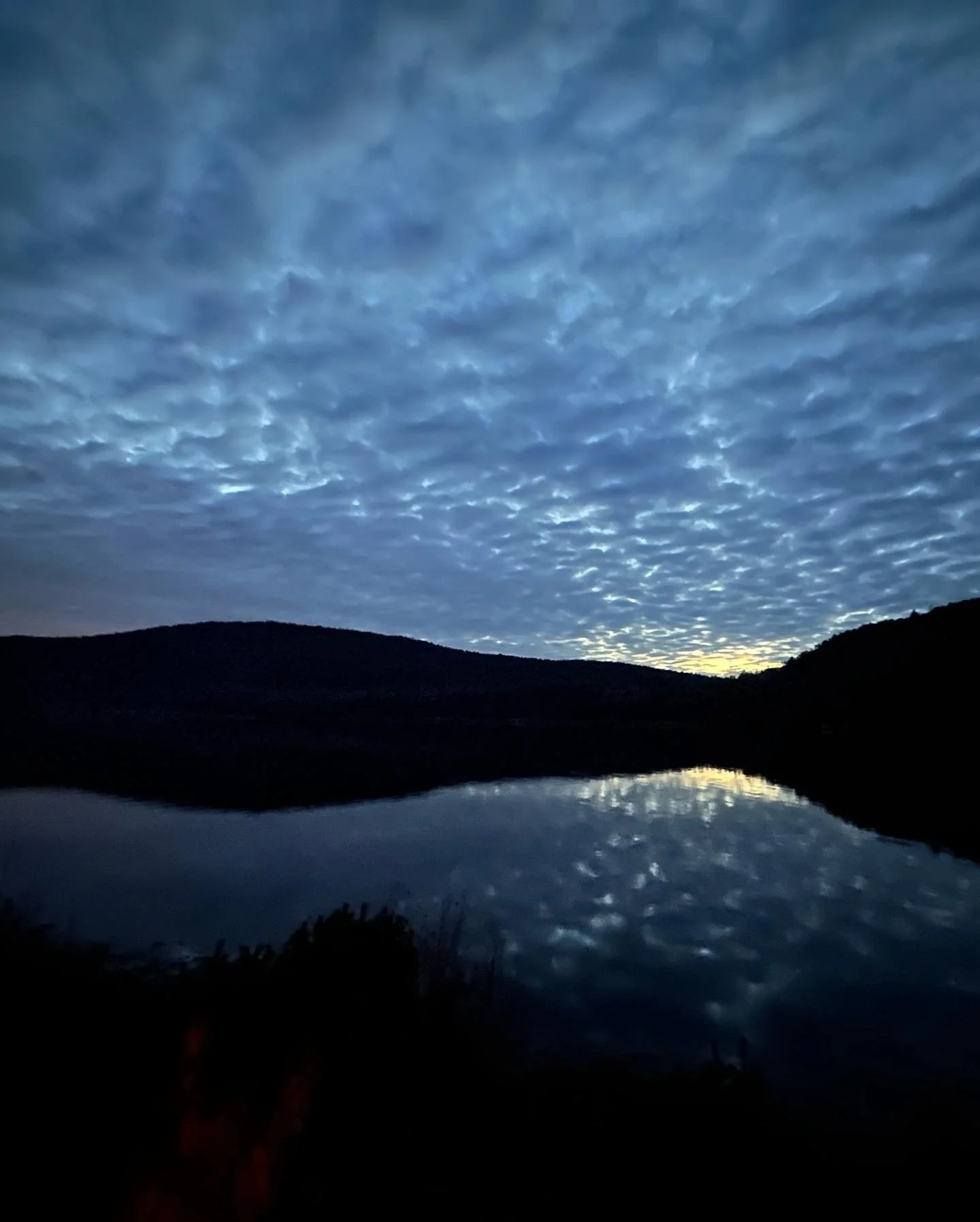 A peaceful scene of a calm lake with a reflection of the dark hills and cloudy sky during dusk or dawn.