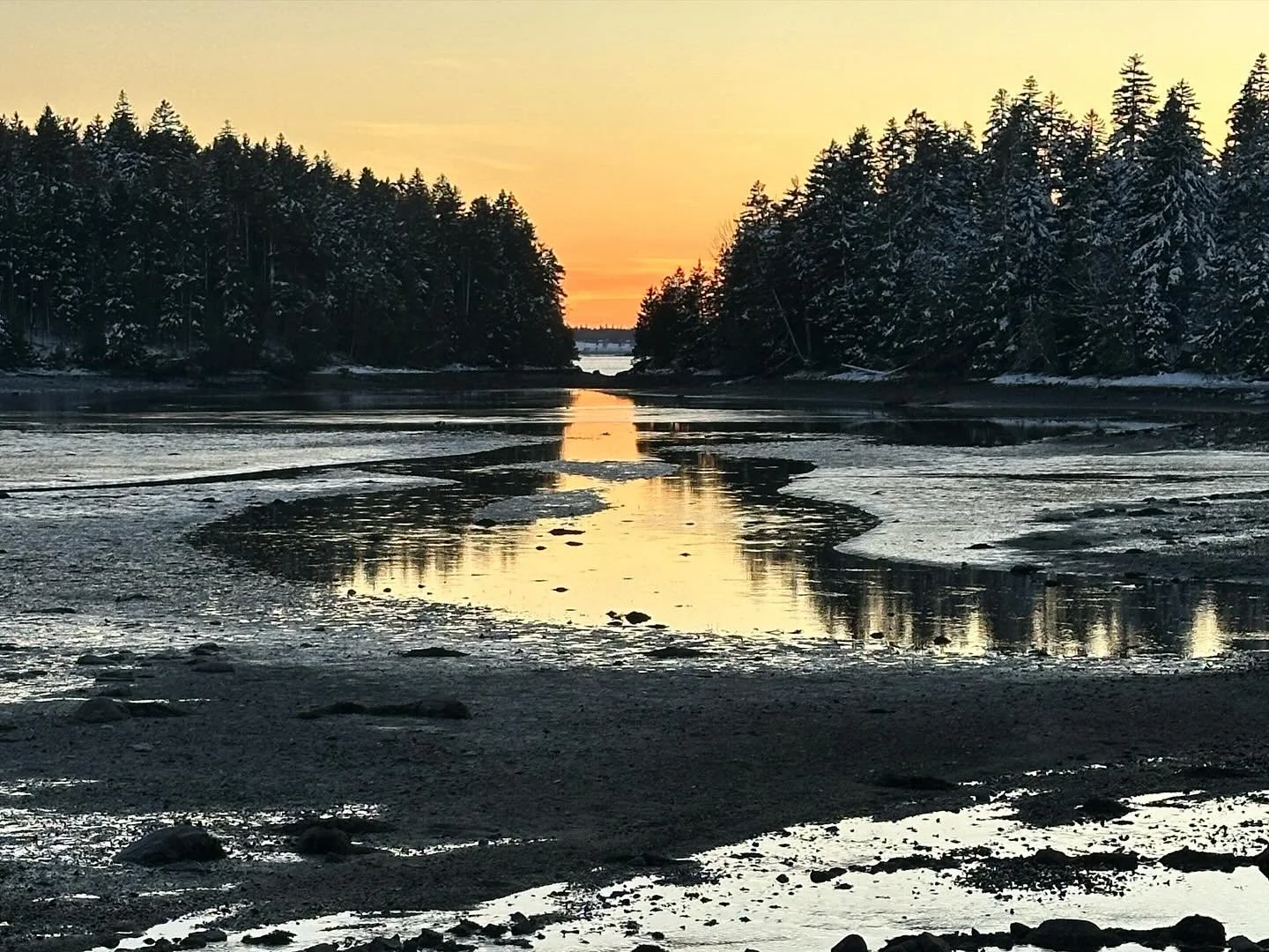 Sunset over a partially frozen river, with snow-covered trees lining the banks on both sides.