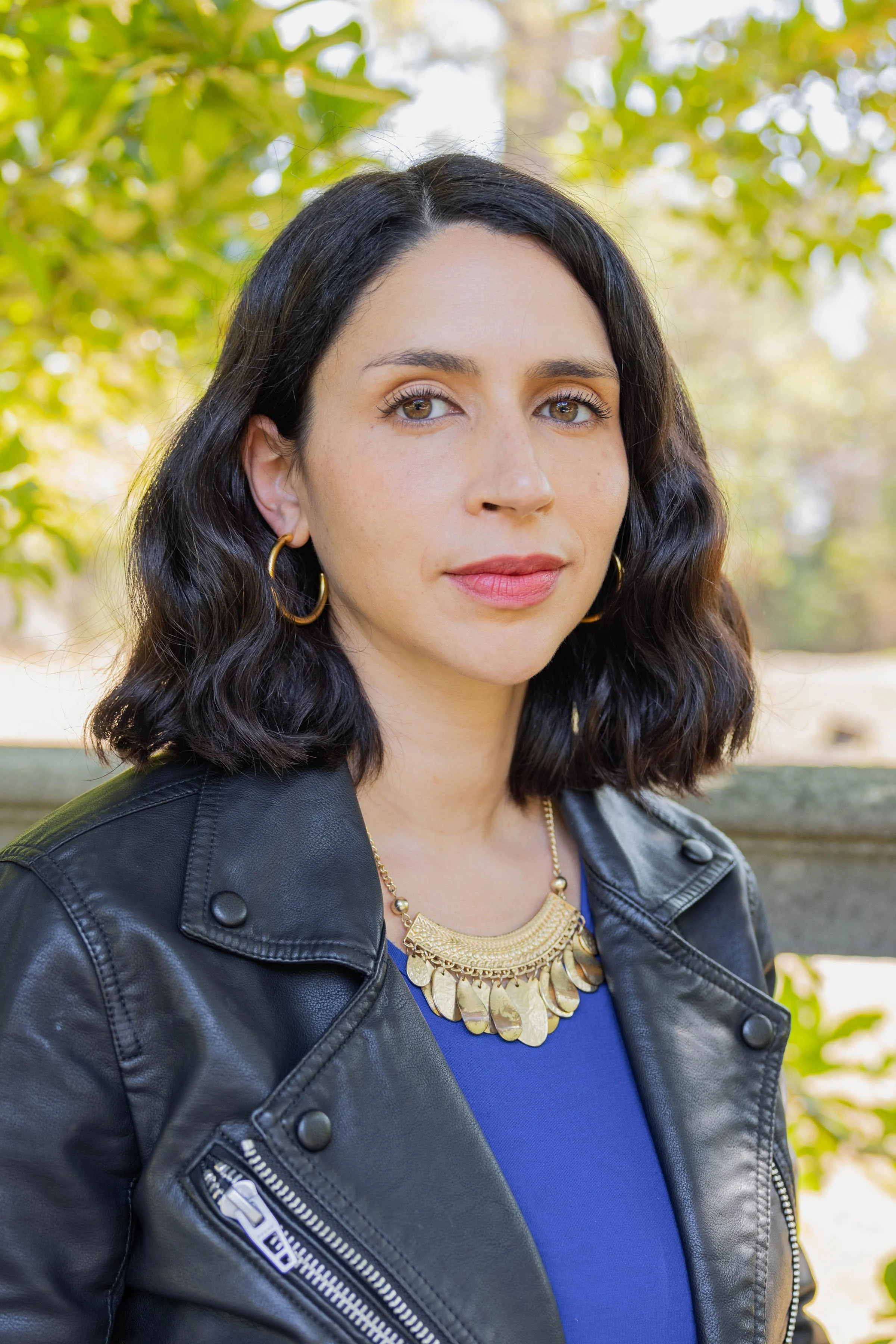 A woman with shoulder-length hair wearing a black leather jacket looks into the camera against a backdrop of leafy trees