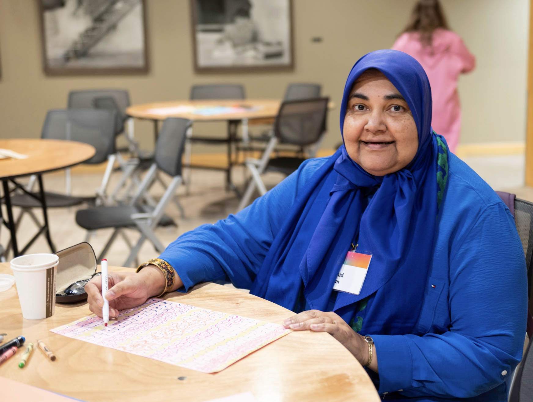 A woman wearing Hijab and a blue outfit sitting on a table writing and looking at the camera