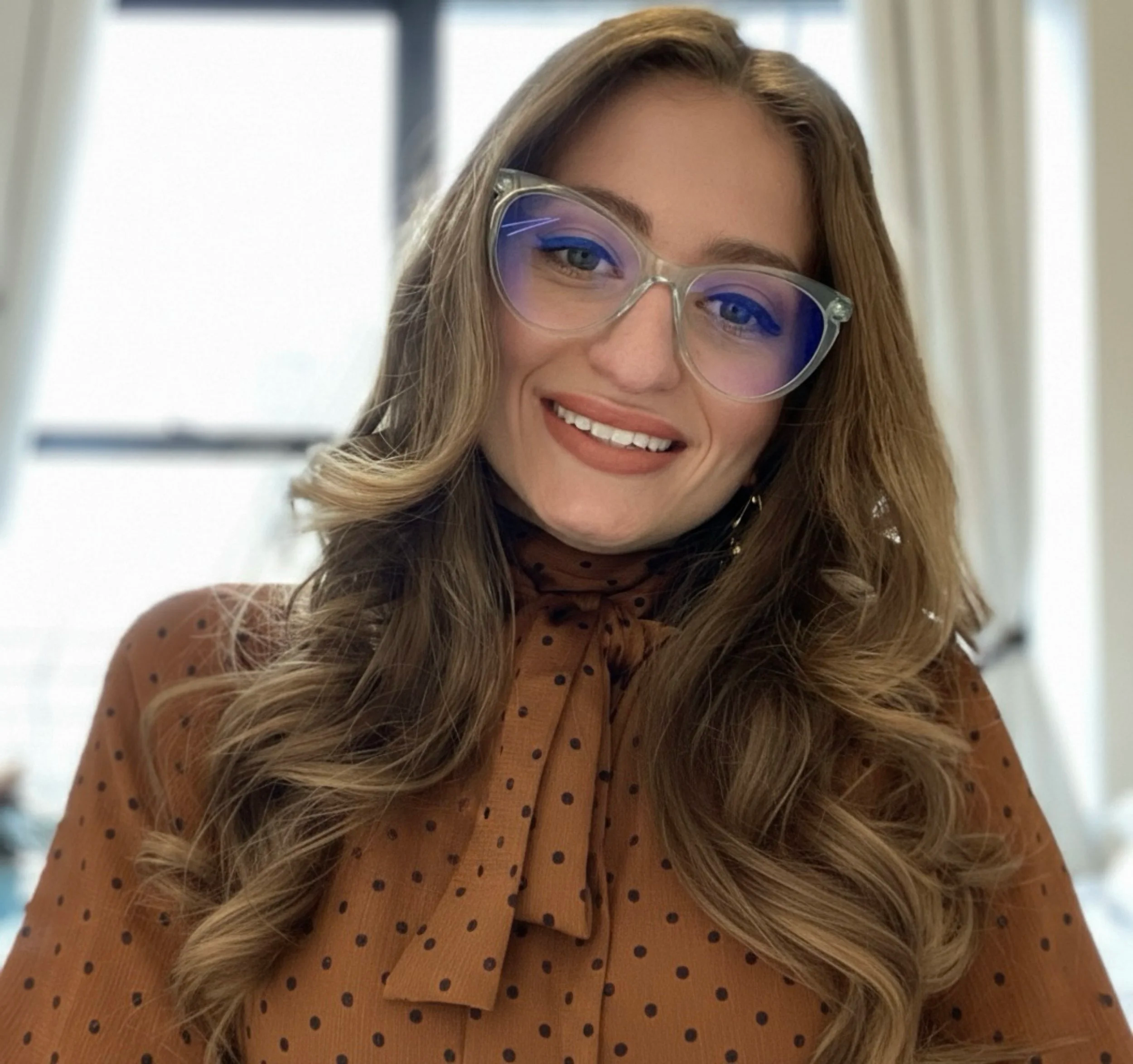 A woman with long hair and large glasses and a polka dot blouse smiles at the camera