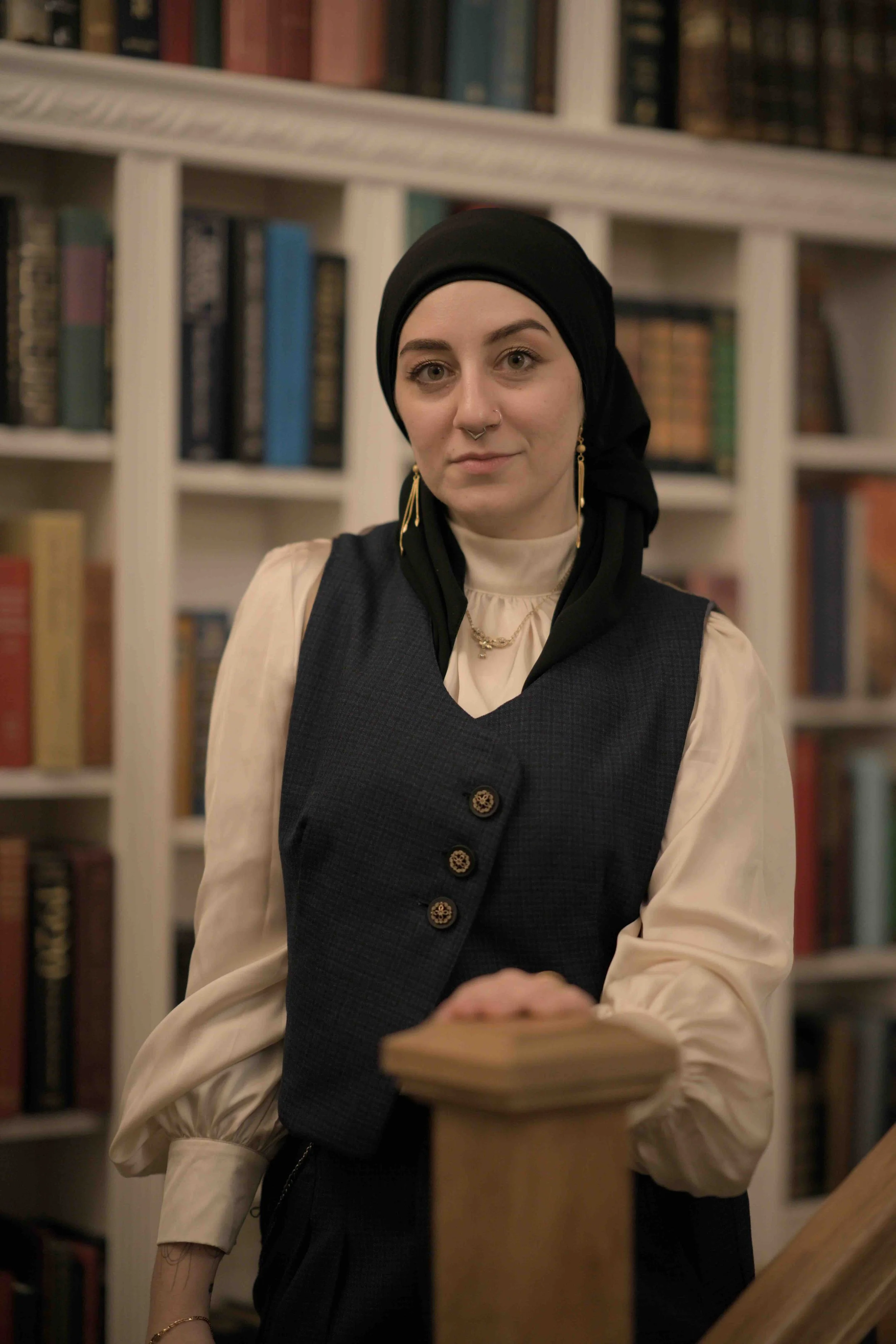 A light-skinned woman in a black hijab and loose-sleeve white blouse and vest looks into the camera in front of a bookshelf