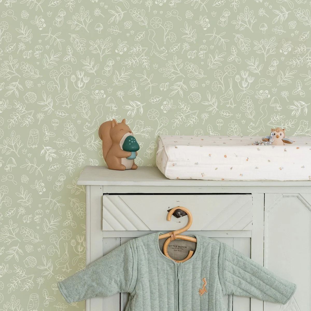 Nursery room with green wallpaper featuring white line drawings of animals, plants, and mushrooms. On top of a white dresser, there is a bear-shaped lamp and a small stuffed animal. A hanging gray baby outfit with an embroidered animal is on a hook below.
