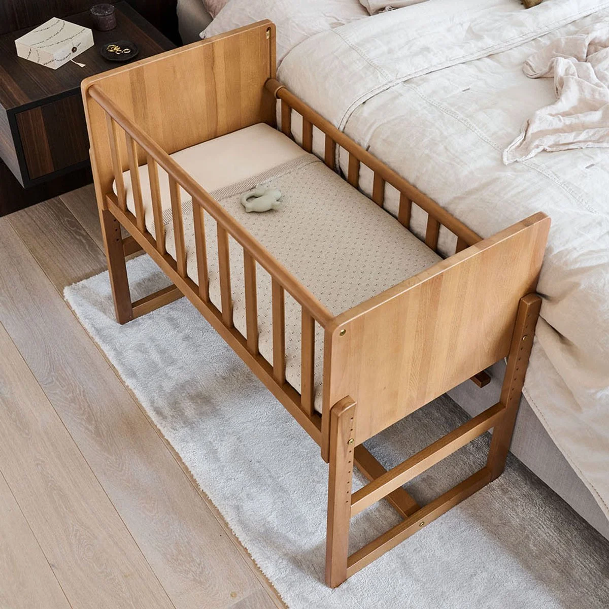 A wooden baby crib next to a bed with white bedding in a bedroom. A small plush toy is on the crib's mattress.