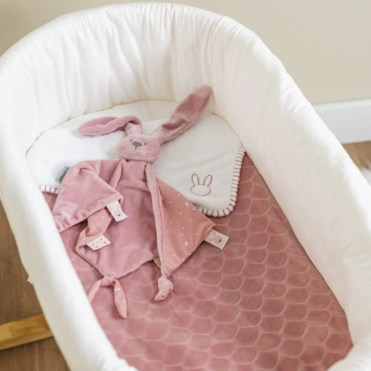 A pink stuffed bunny toy with long ears lying inside a cream-colored baby bassinet with a pink textured mattress pad, a white burp cloth with a bunny embroidery, and a pink blanket with tied knots at the ends.