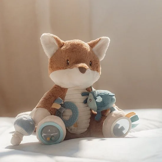 A plush fox stuffed animal with wheels and a small stuffed fish is sitting on a white bed. The background is a plain beige wall.