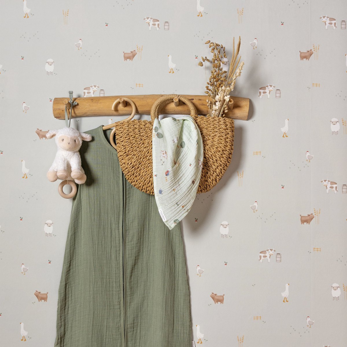 Baby clothes and accessories hanging on a wooden coat rack against a farm animal themed wallpaper. Items include a stuffed lamb toy, a green dress, a cloth with small illustrations, and dried plants.