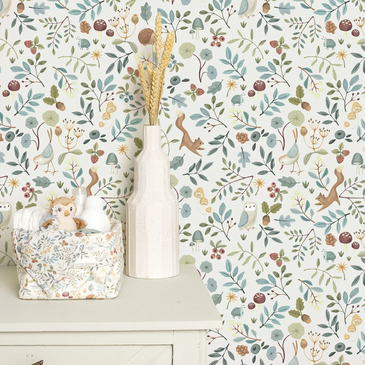 Decorative nursery wall with woodland-themed wallpaper featuring trees, mushrooms, owls, birds, squirrels, and berries, arranged behind a white dresser with a plush lion toy, a fabric storage basket with a plush toy, and a tall white vase with dried wheat stalks.