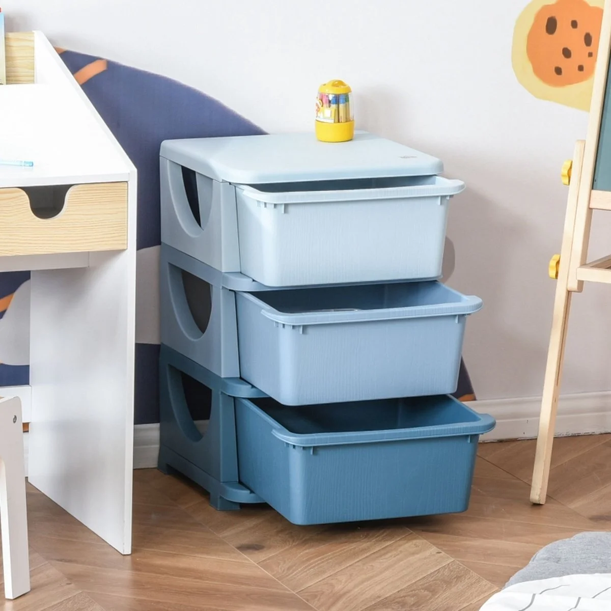A stack of three grey plastic storage bins with open tops, placed on a wooden floor in a room with a white wall. A yellow toy with colorful parts is on top of the bins. To the left, part of a white desk with a drawer, and to the right, a wooden easel with yellow knobs. Part of a wall decoration with a cookie illustration is visible.
