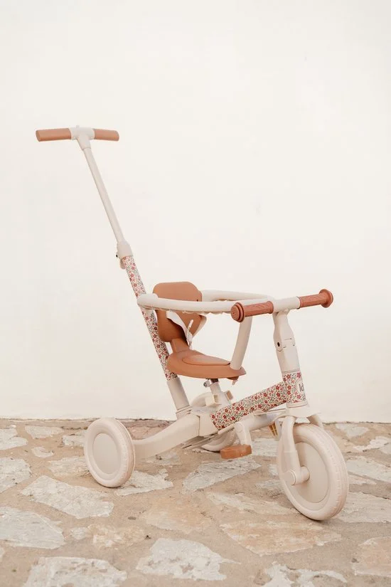 A small children's tricycle with a white frame, light brown seat, and handlebar, decorated with a floral pattern on the frame, placed against a plain white wall on a stone floor.