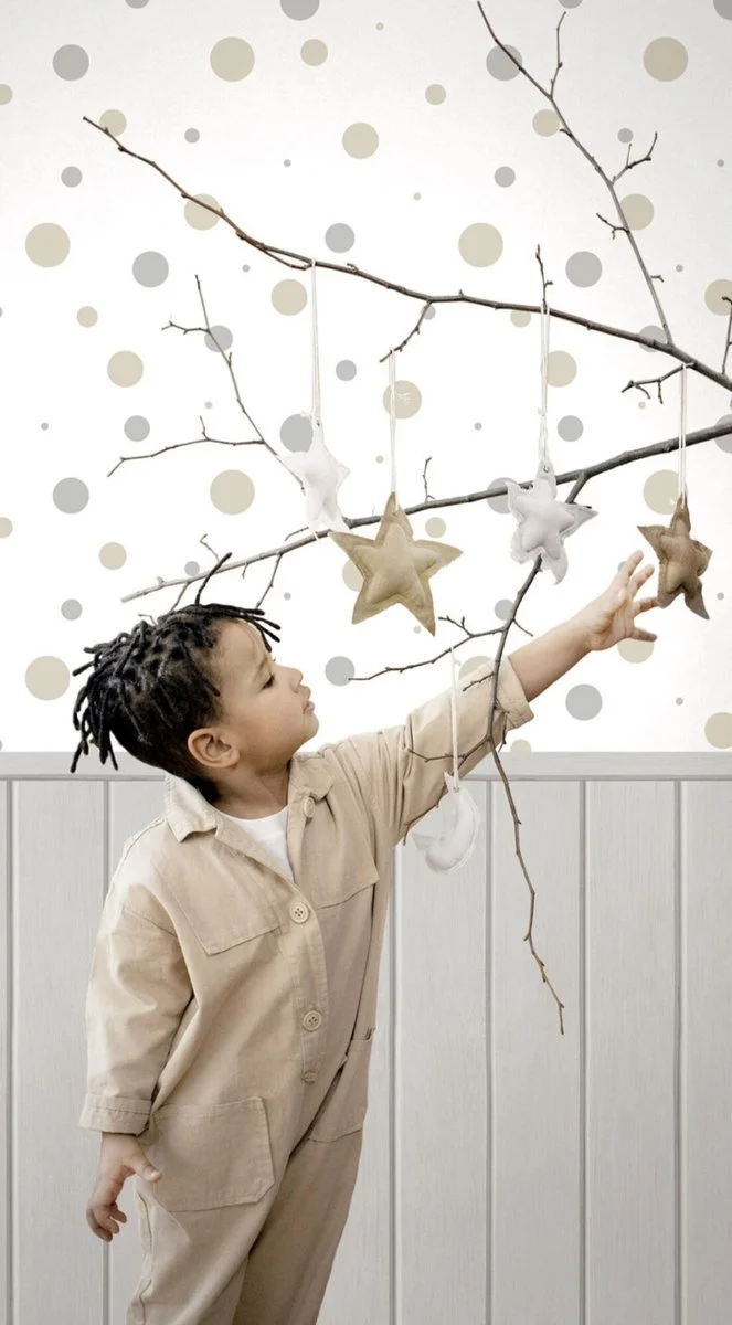 A young boy with braided hair reaching up towards hanging star-shaped ornaments on a branch in front of a wall with polka dot wallpaper.